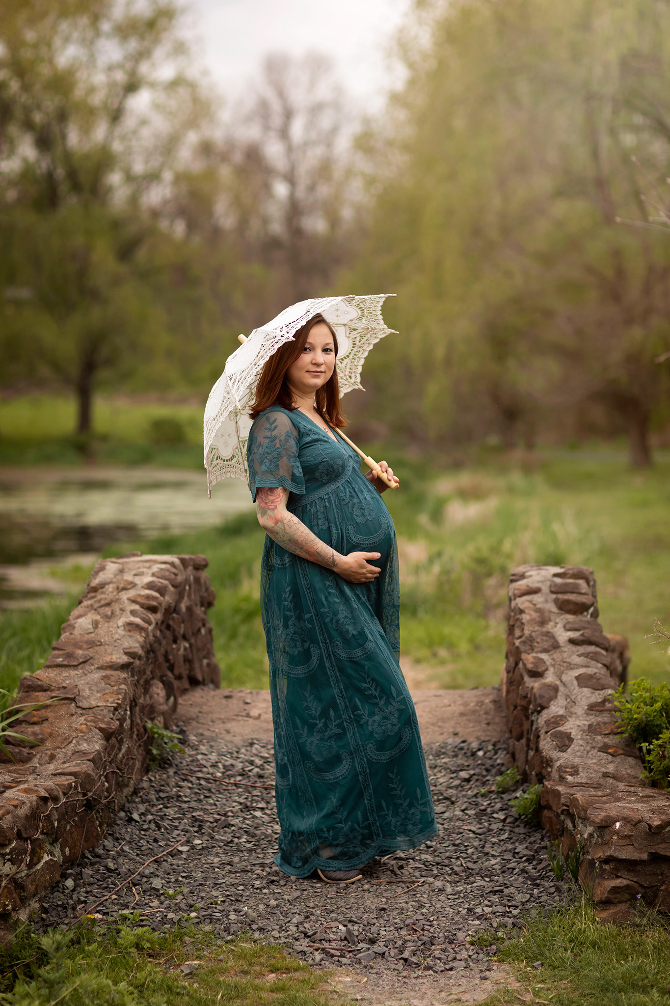 Expecting mom holds her belly and a white lace umbrella while standing on a bridge I Prenatal Massage near Morristown NJ