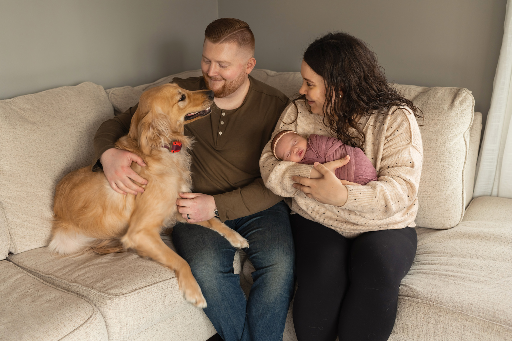 A golden retriever smiles while sitting on a couch with mom and dad with their newborn baby girl