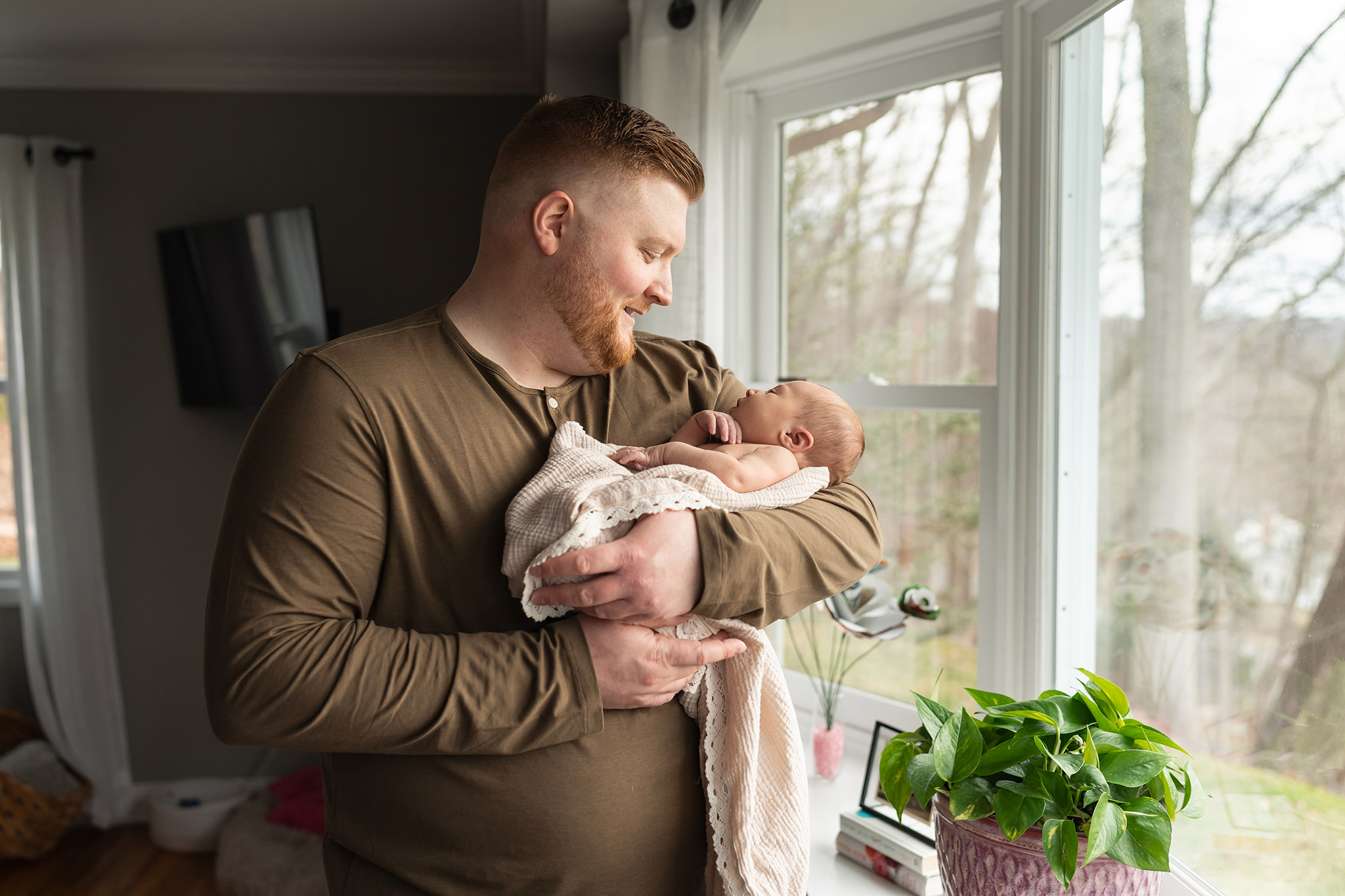 A happy father smiles down at his newborn baby in his arms while standing in a window thanks to a 3d ultrasound northern nj