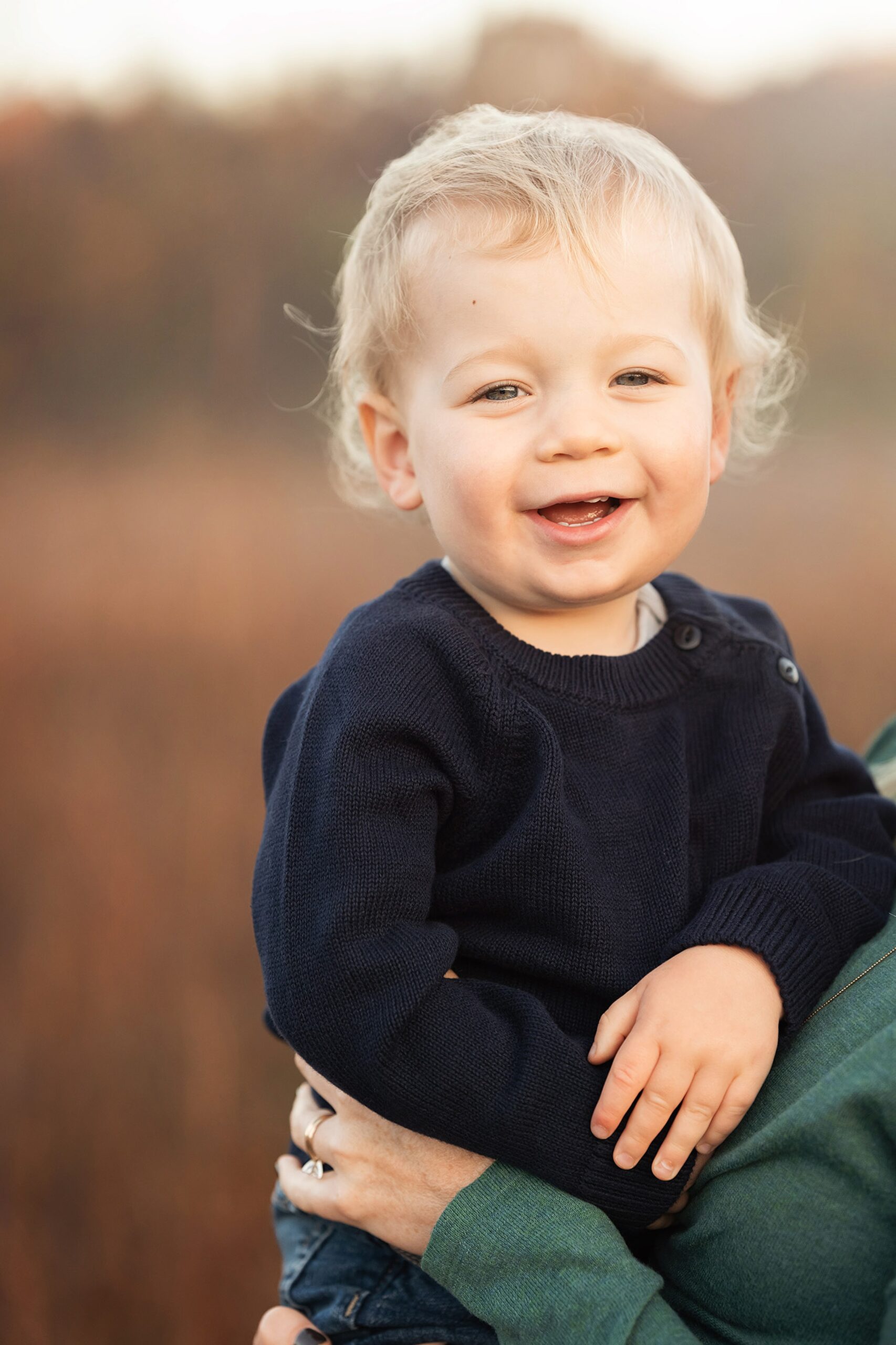 A happy toddler boy sits in mom's arms while smiling in a park in a blue sweater