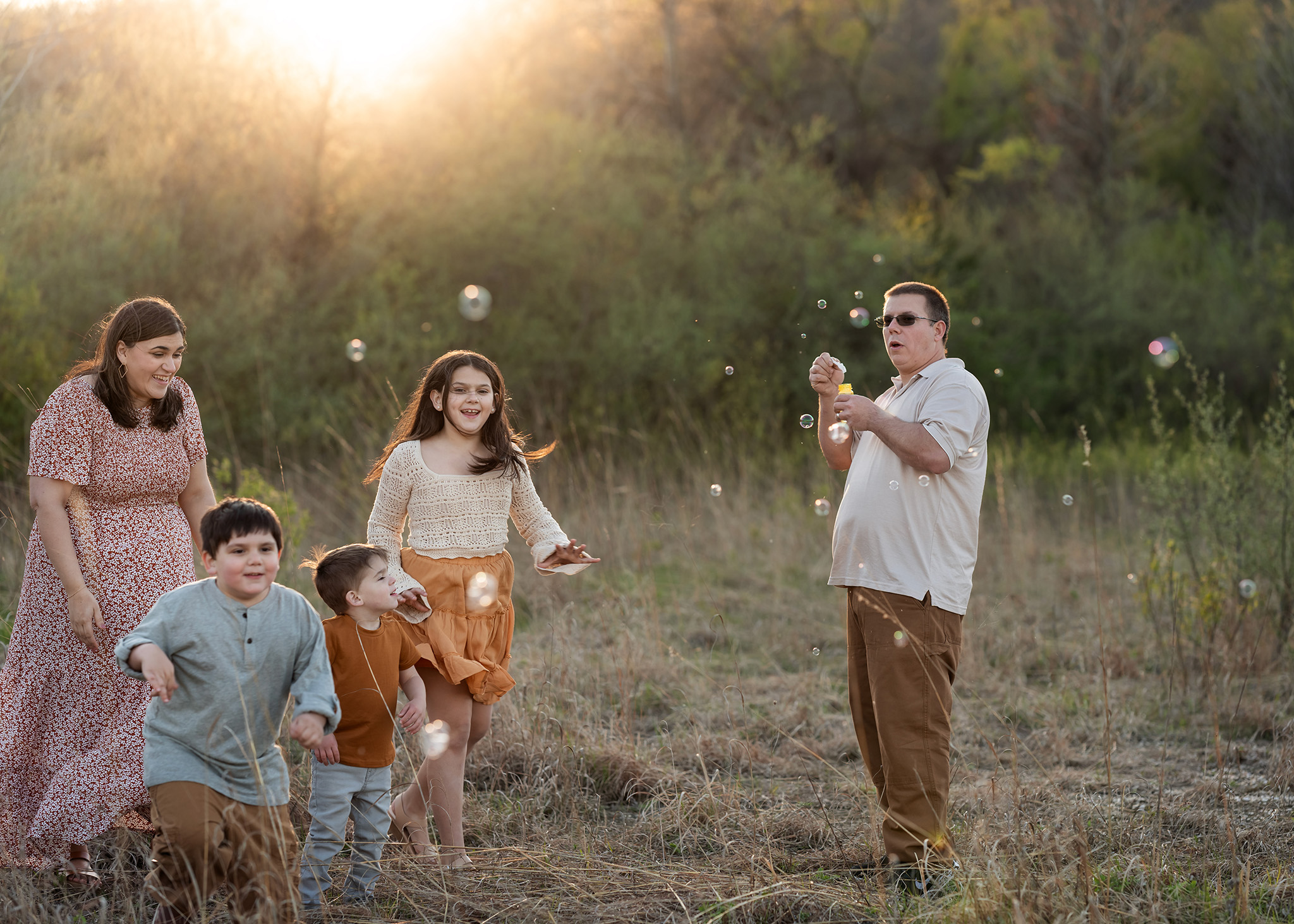 A dad blows bubbles for his three children to chase in a park at sunset with mom after meeting babysitters nj