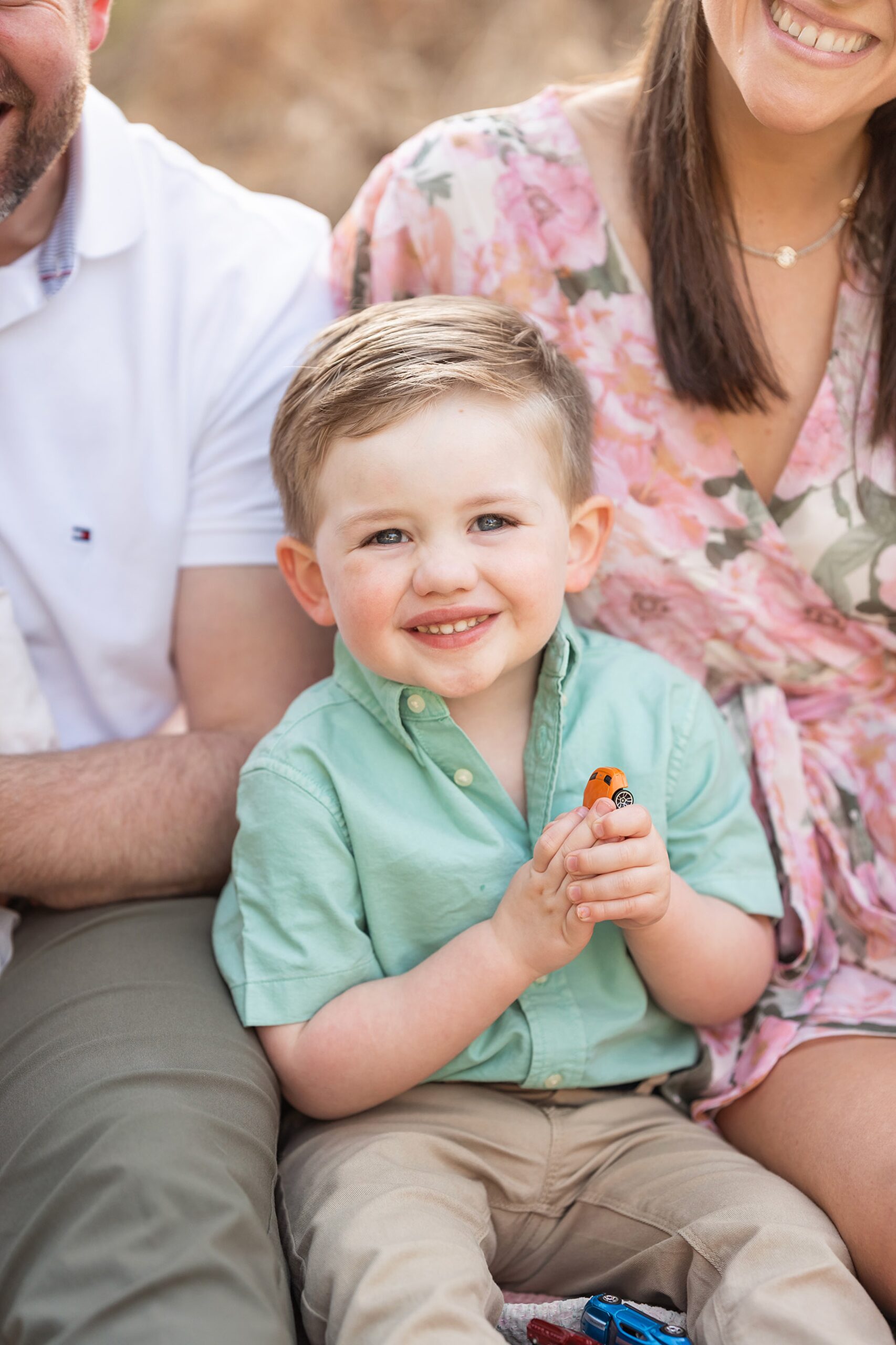 A happy toddler boy plays with toy cars while sitting in a park between mom and dad