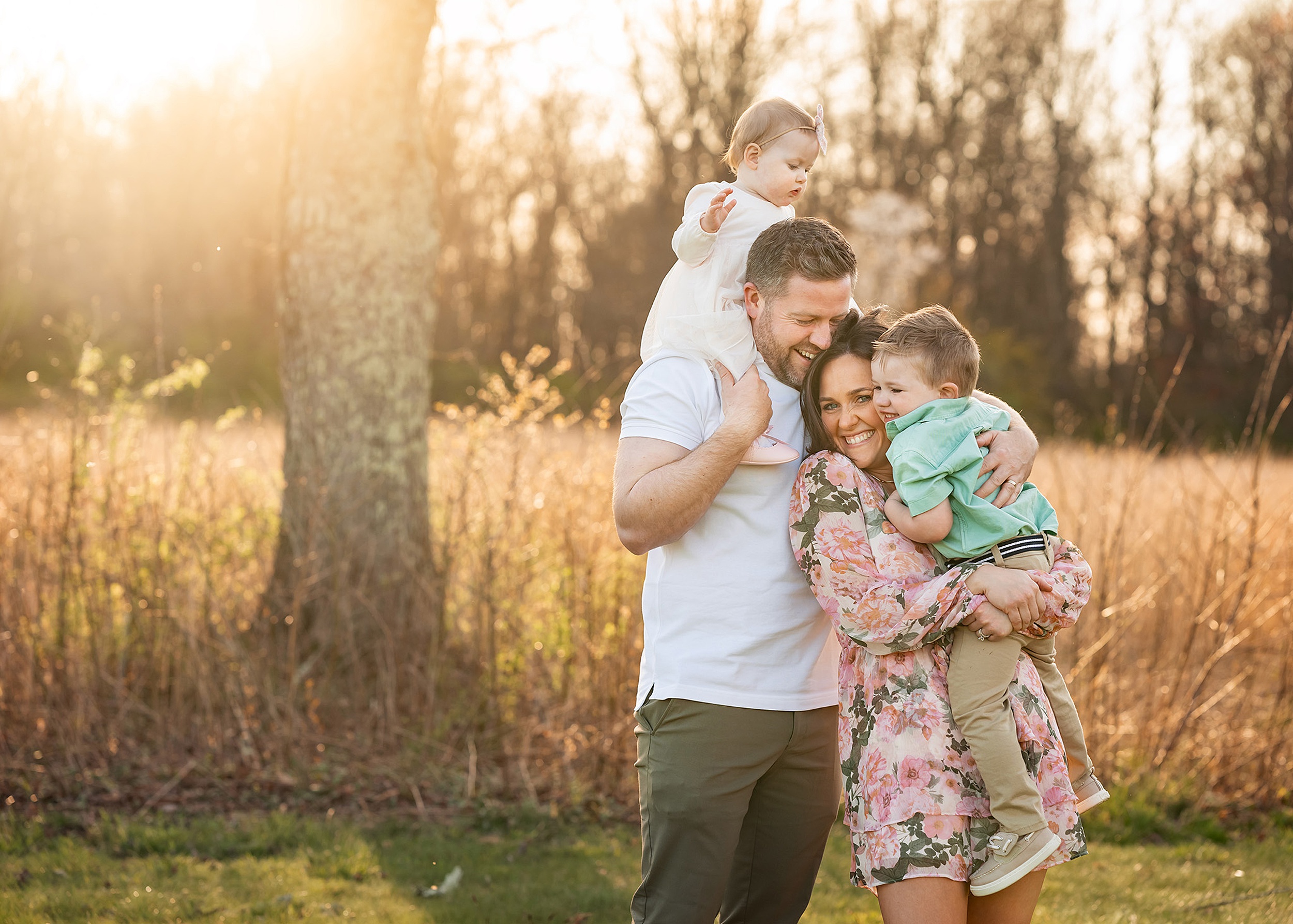Happy mom and dad stand and laugh together while holding their toddler son and daughter in a park at sunset after visiting daycares in morris county nj