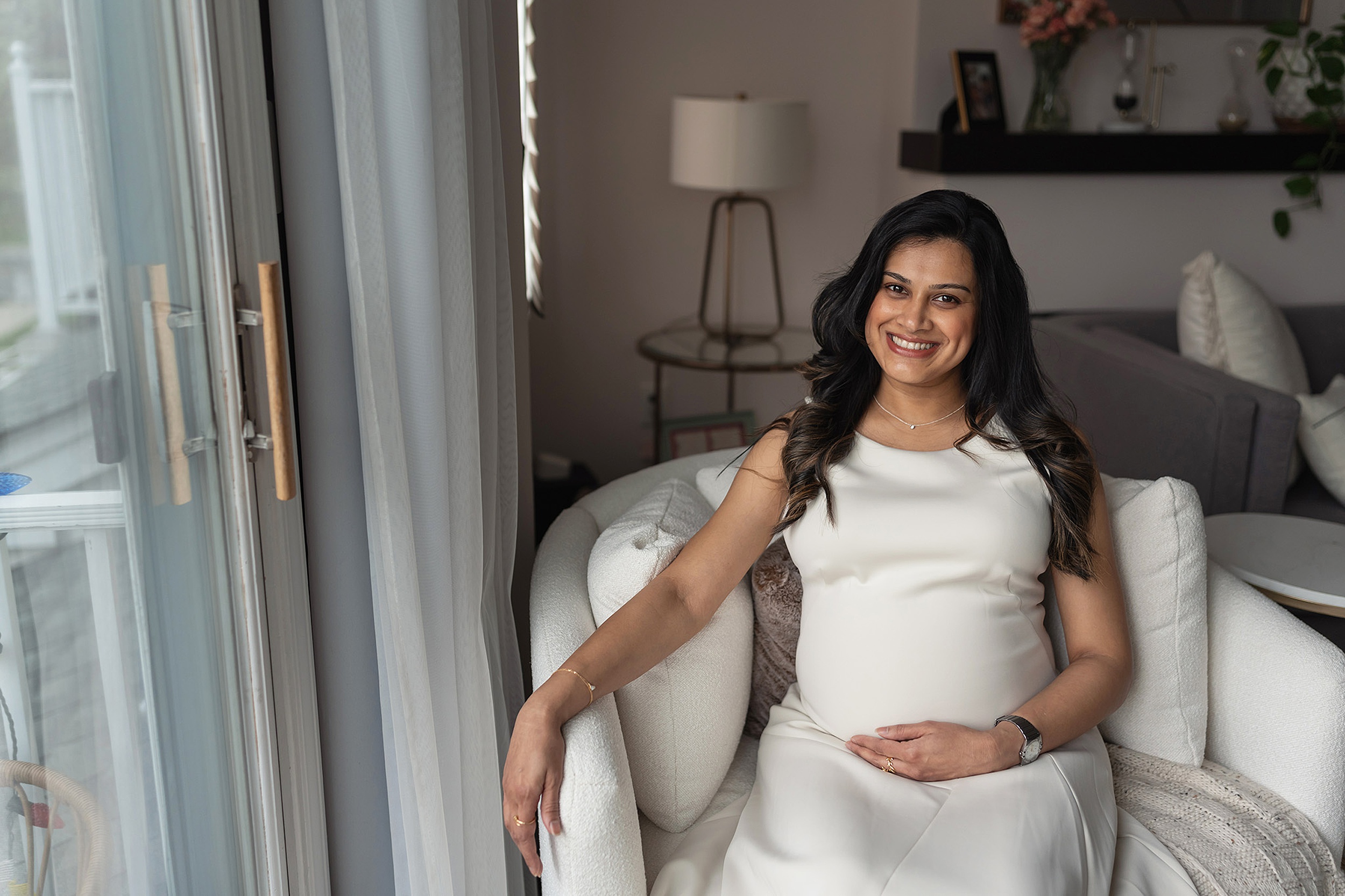 A smiling mother to be holds her bump while sitting in a window in a white maternity gown