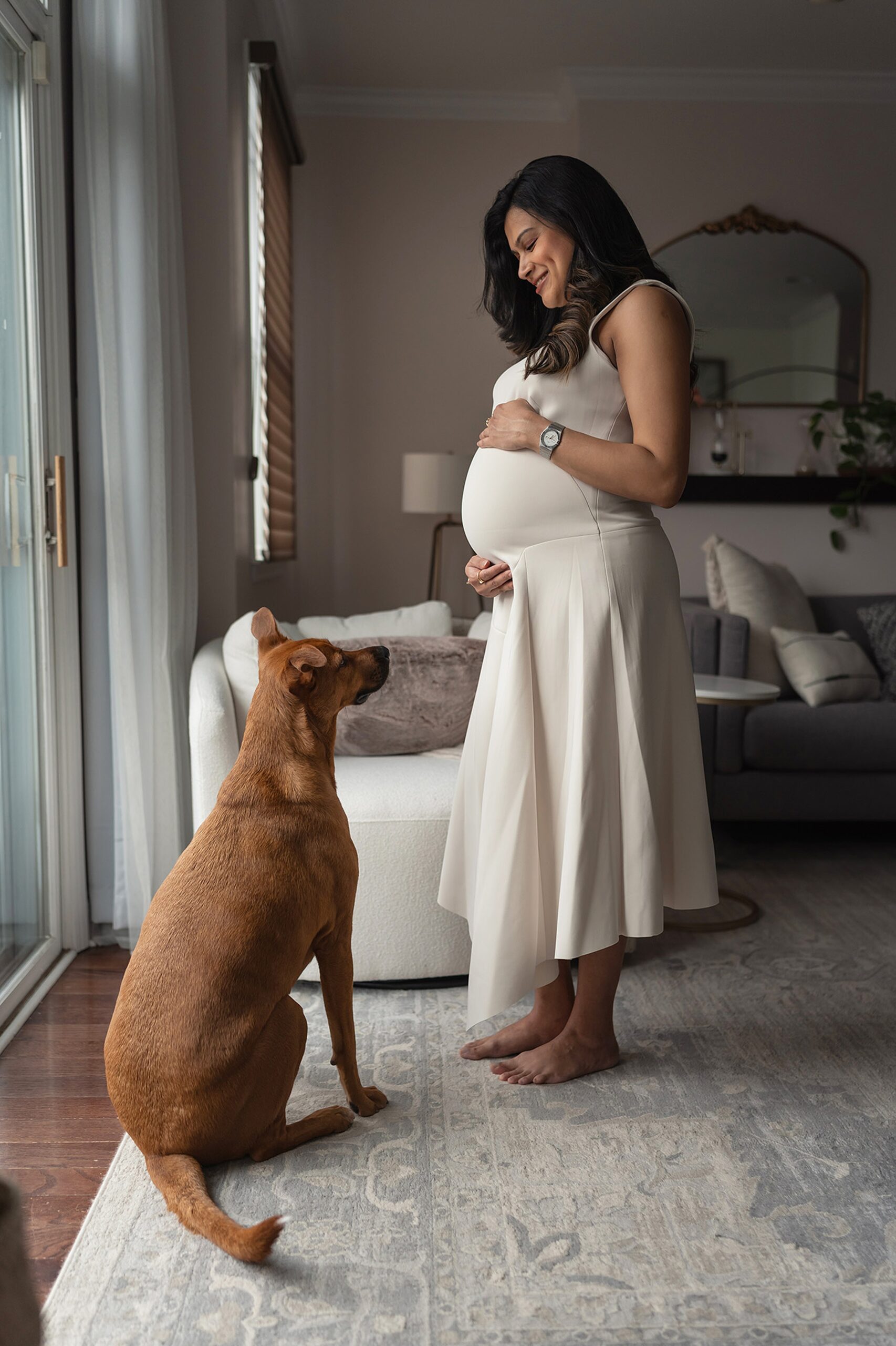 A mom to be in a white maternity gown stands in her living room smiling down to her dog in front of her after visiting doulas in nj