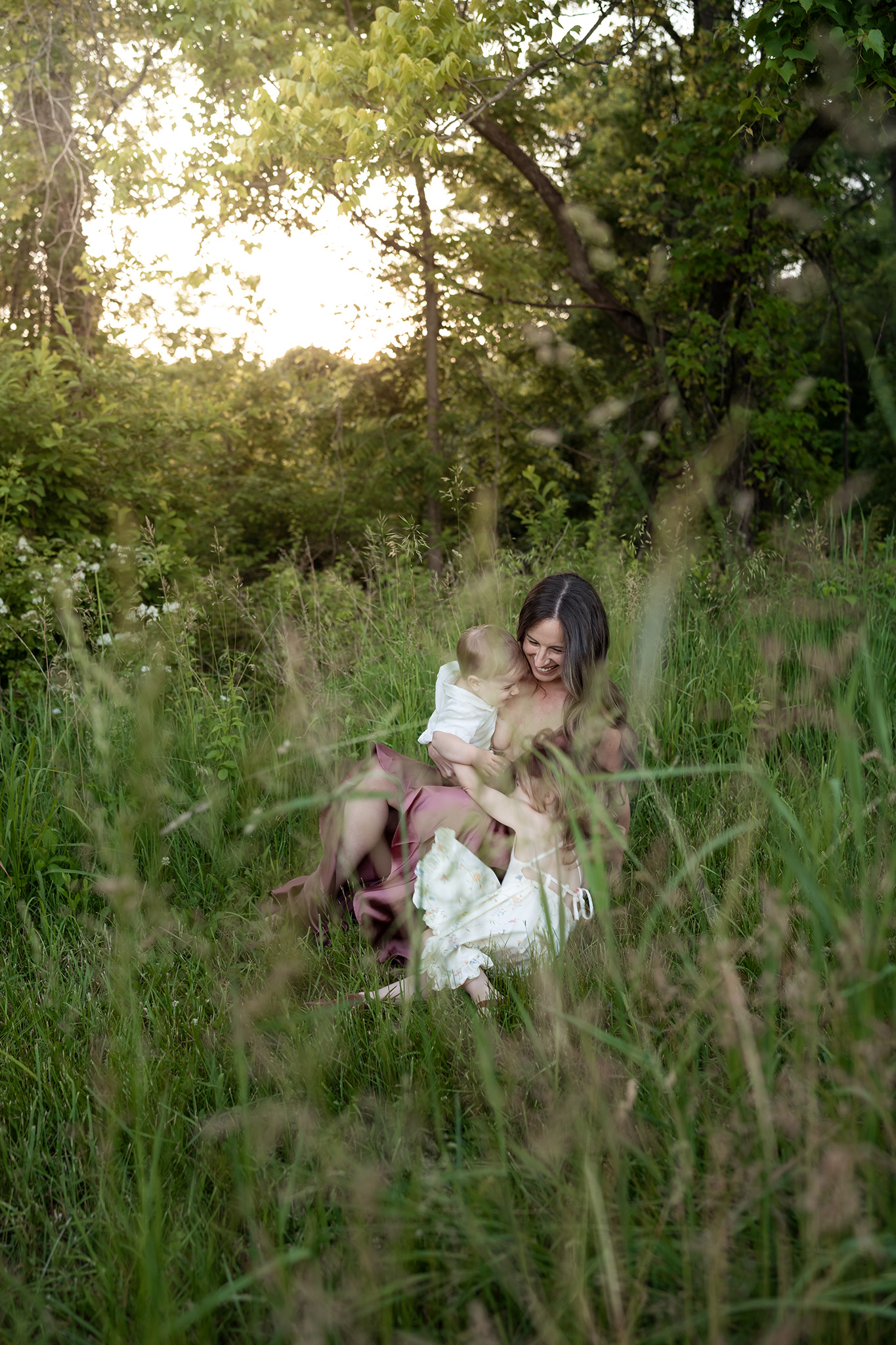 A mother plays with her toddler son and daughter while sitting in some tall grass at sunset