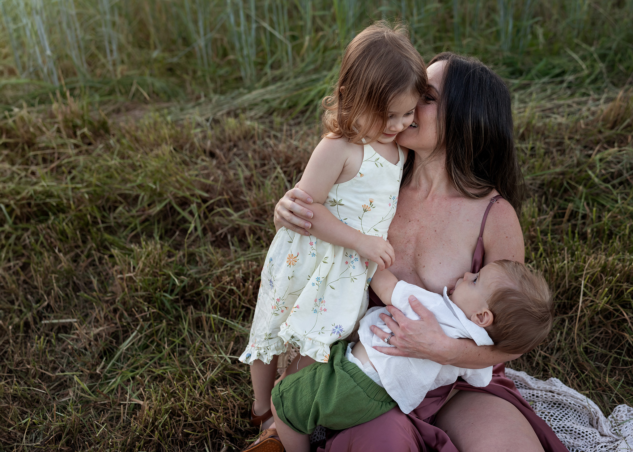 A mother breastfeeds her toddler son in a park while her toddler daughter gets a hug and kiss thanks to fertility clinics new jersey