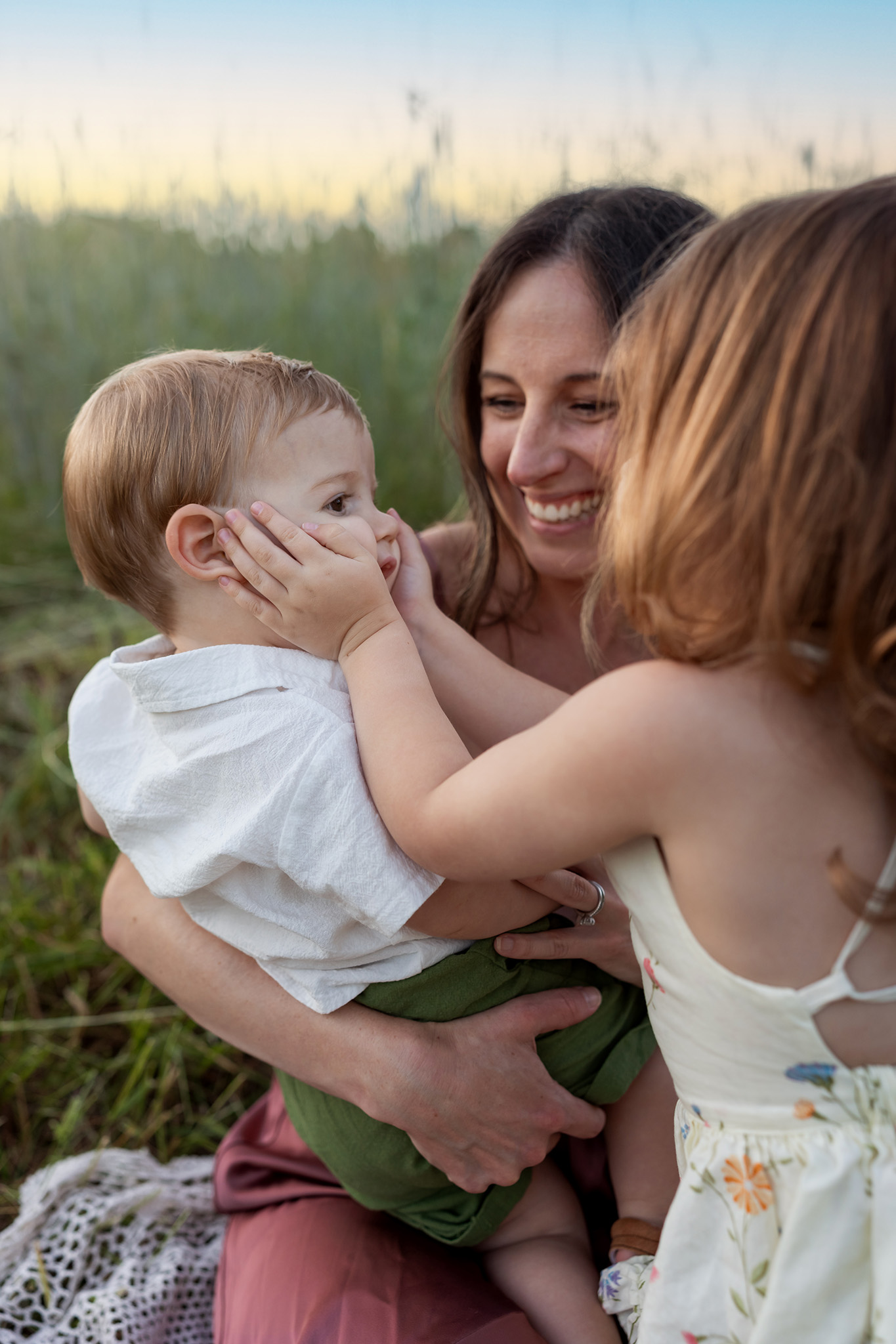 A happy mom holds her toddler son while her toddler daughter squishes his face in a park field of tall grass thanks to fertility clinics new jersey