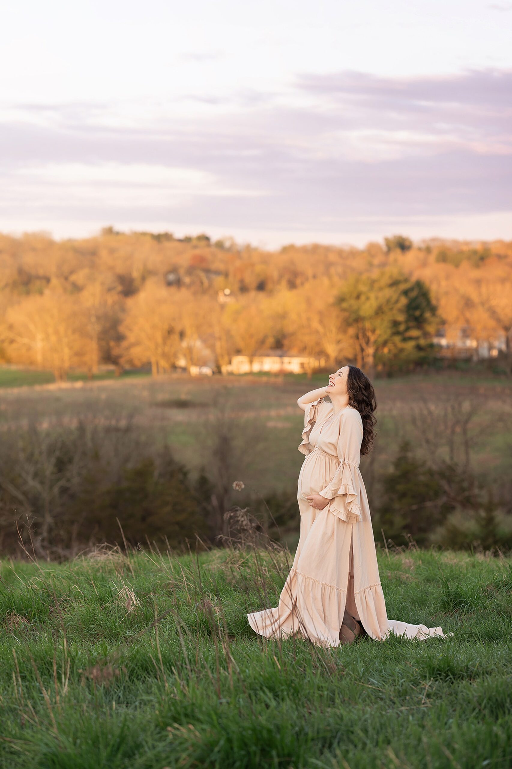 A laughing mother to be in a cream maternity gown laughs up to the sky while walking through a grassy hill at sunset