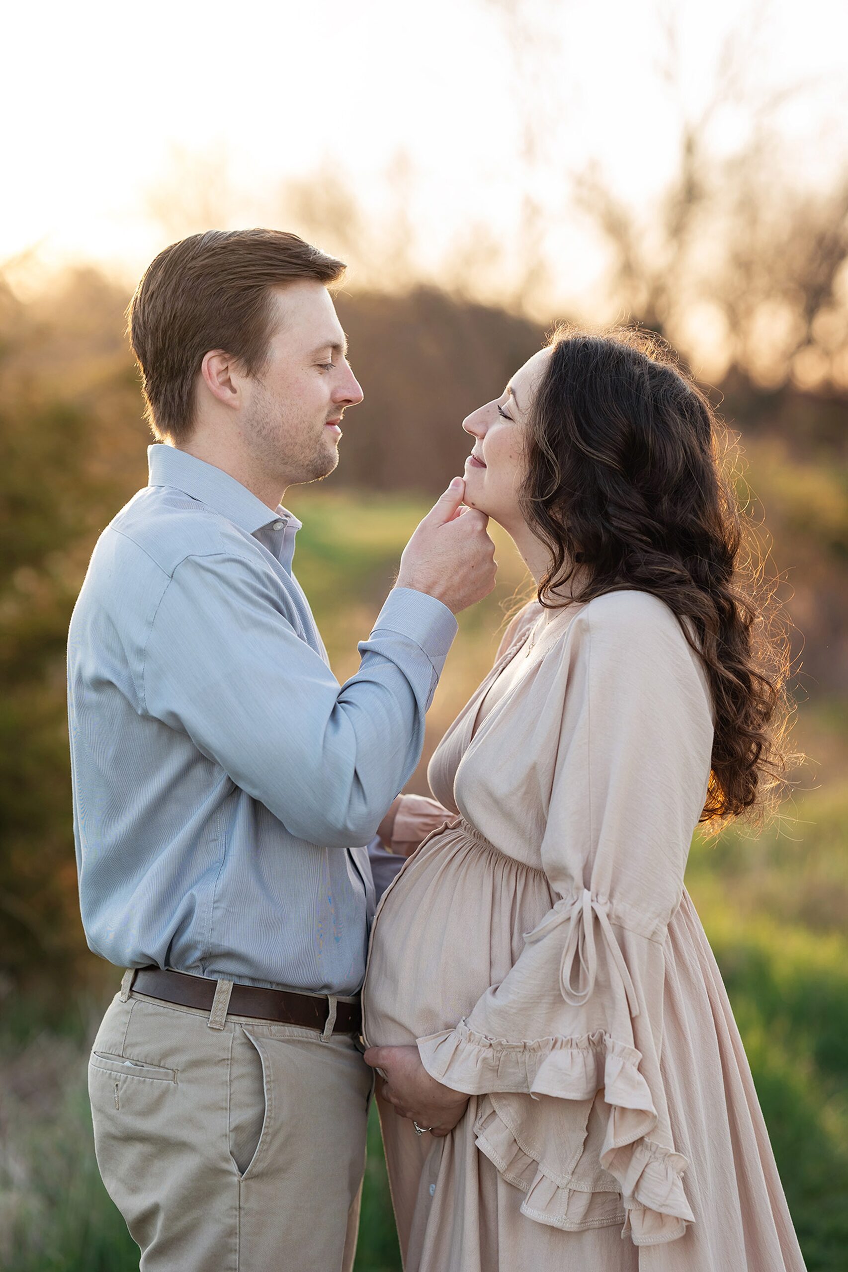 A happy father to be holds his pregnant wife's chin while standing in a park at sunset