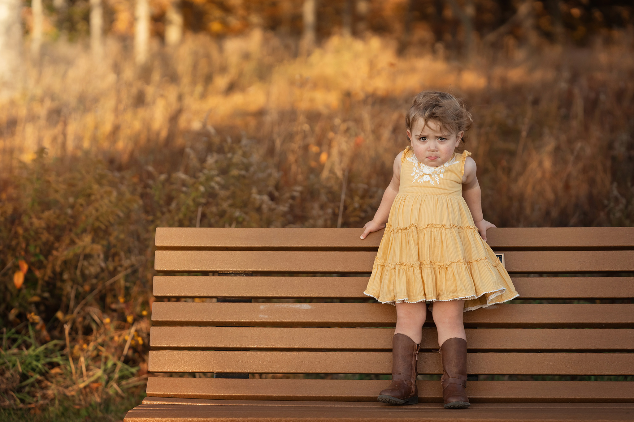 A toddler girl stands on a park bench in a yellow bench and leather boots