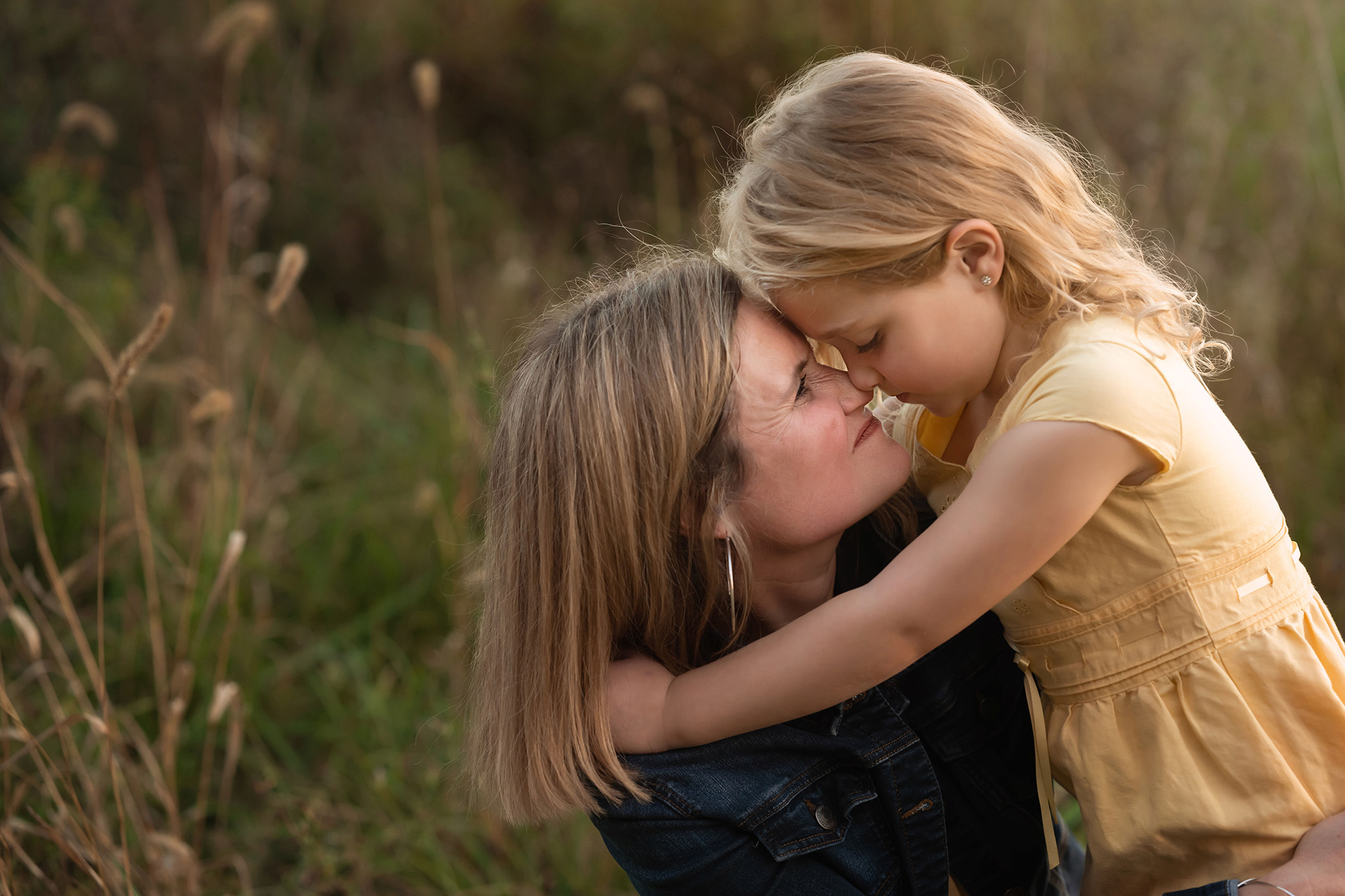 A toddler girl in a yellow dress snuggles her nose to her mom's while in a park at sunset after meeting nannies in nj