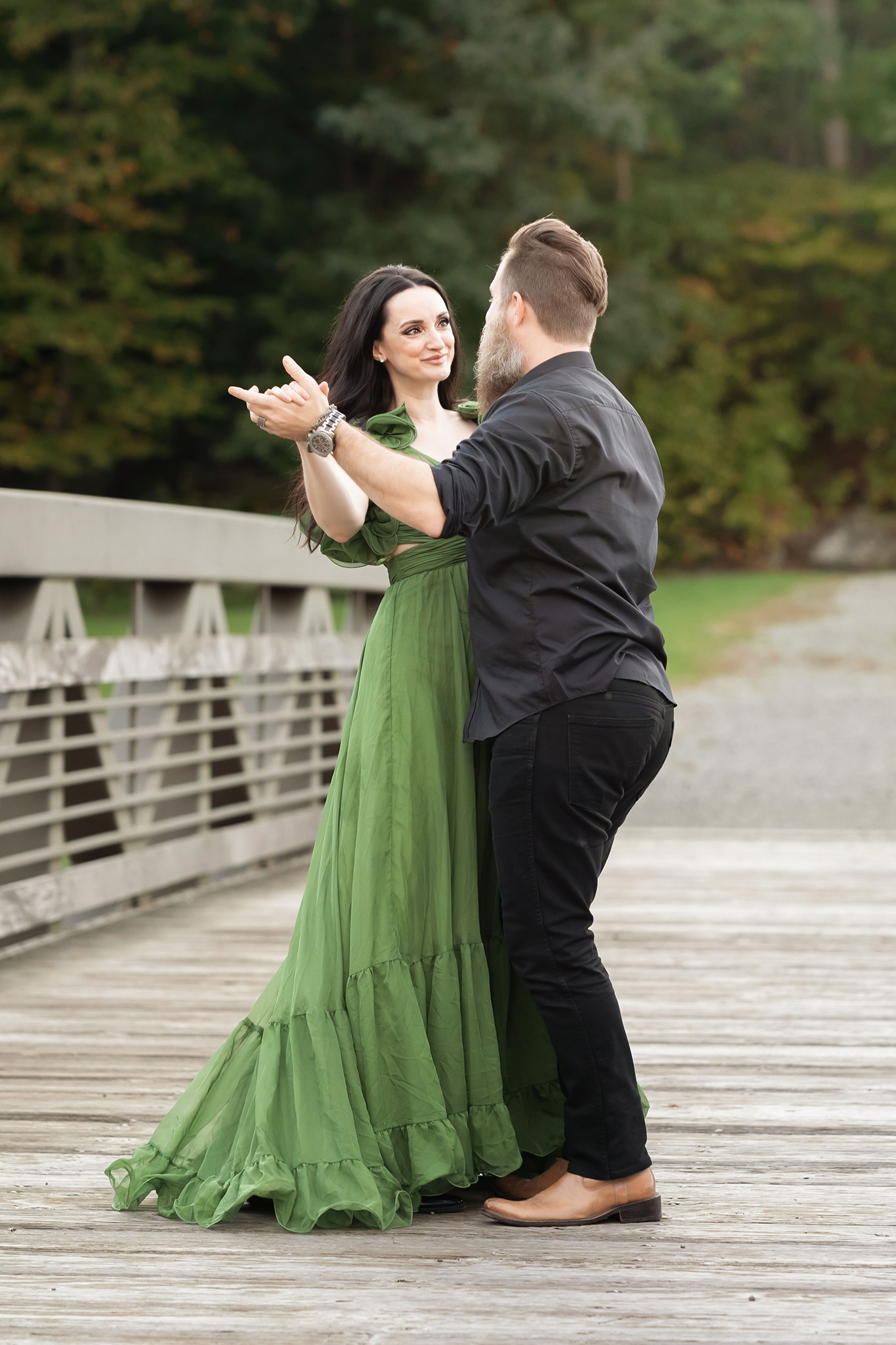 A happy mother in a green dress dances with her husband in black on a park bridge