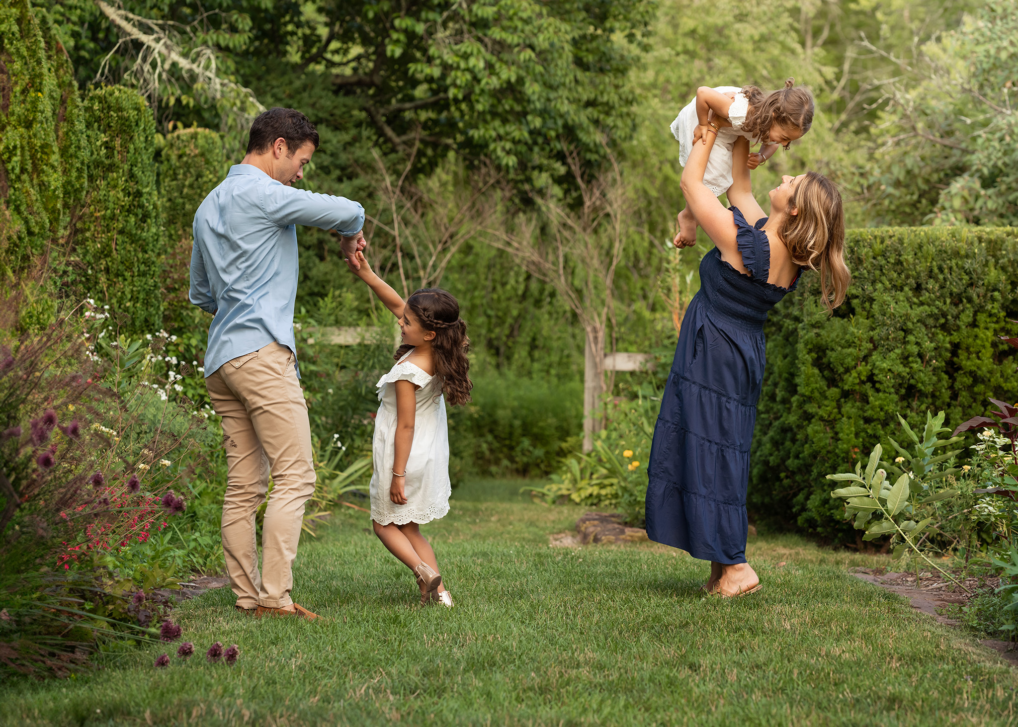 A mom and dad dance and play with their toddler daughters in white dresses in a garden path