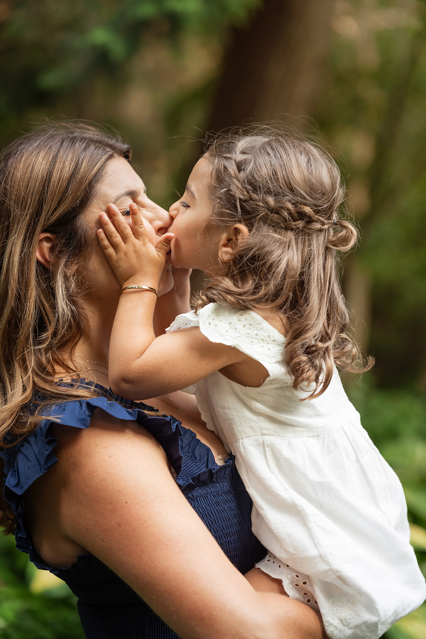 A toddler girl holds mom's face while kissing her in a park in a white dress after visiting children's boutiques nj