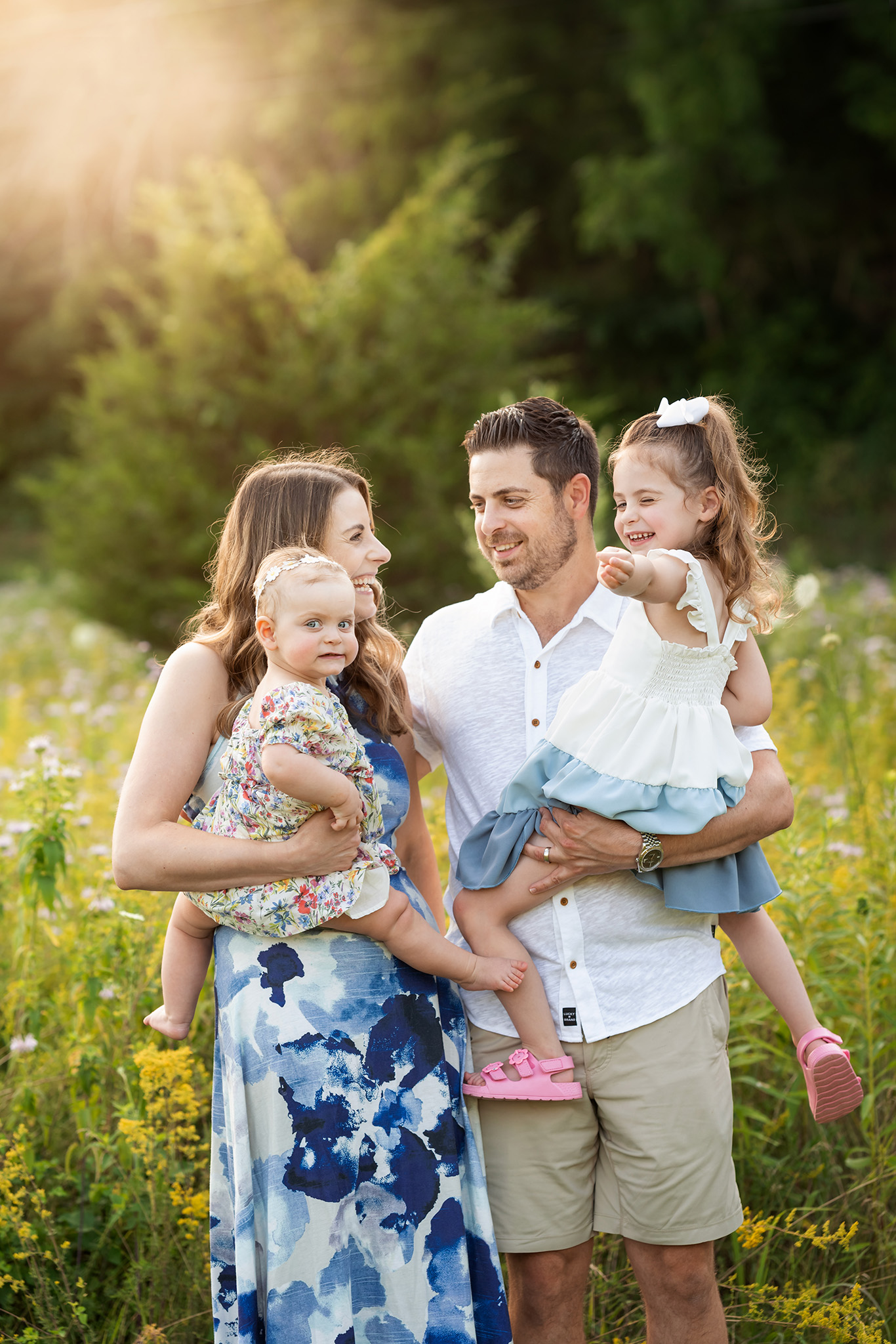 Happy parents giggle with their 2 toddler daughters on their hips in a field of wildflowers after finding a diaper service nj