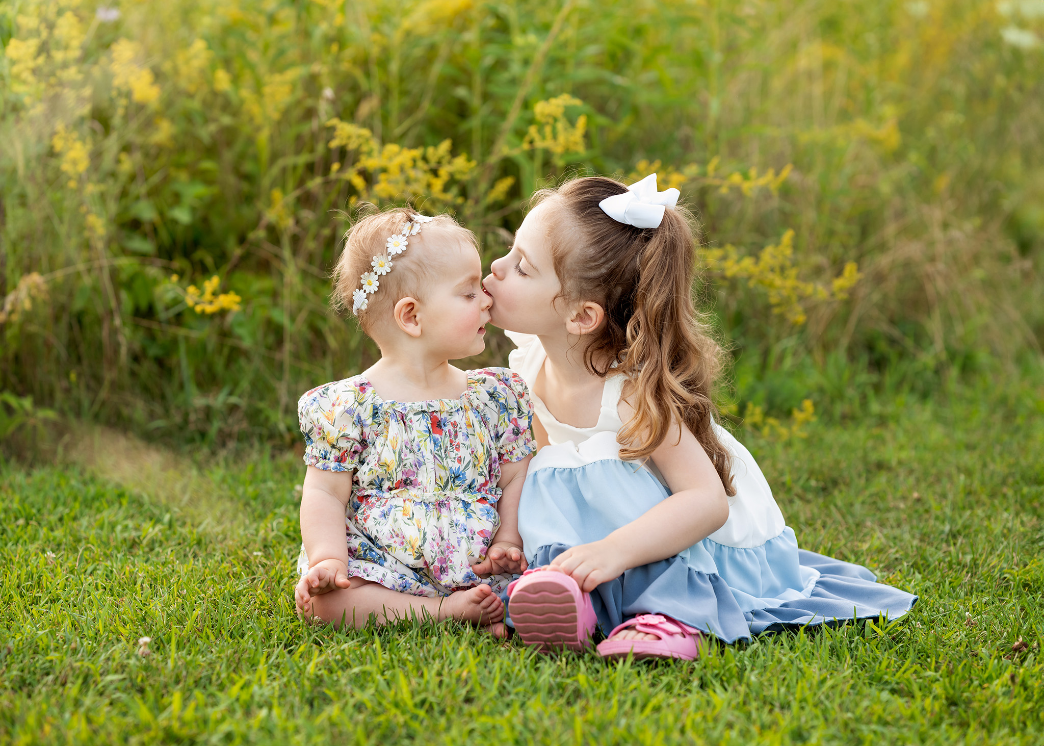 A toddler girl in a blue and white dress kisses her younger sister while sitting in a lawn at sunset