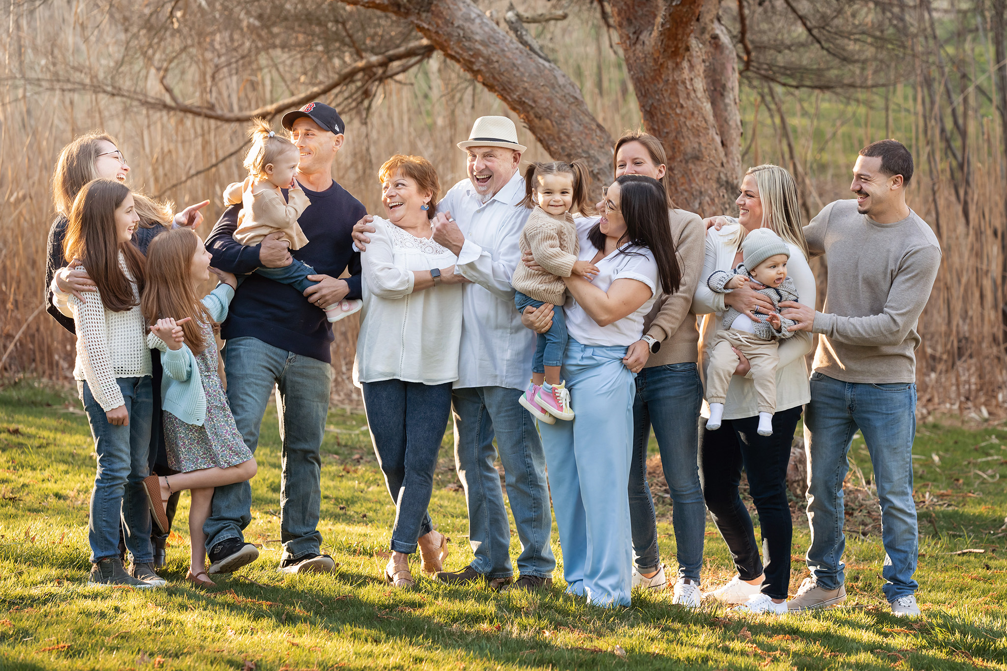 A large extended family laughs together while standing under a tree in a park at sunset