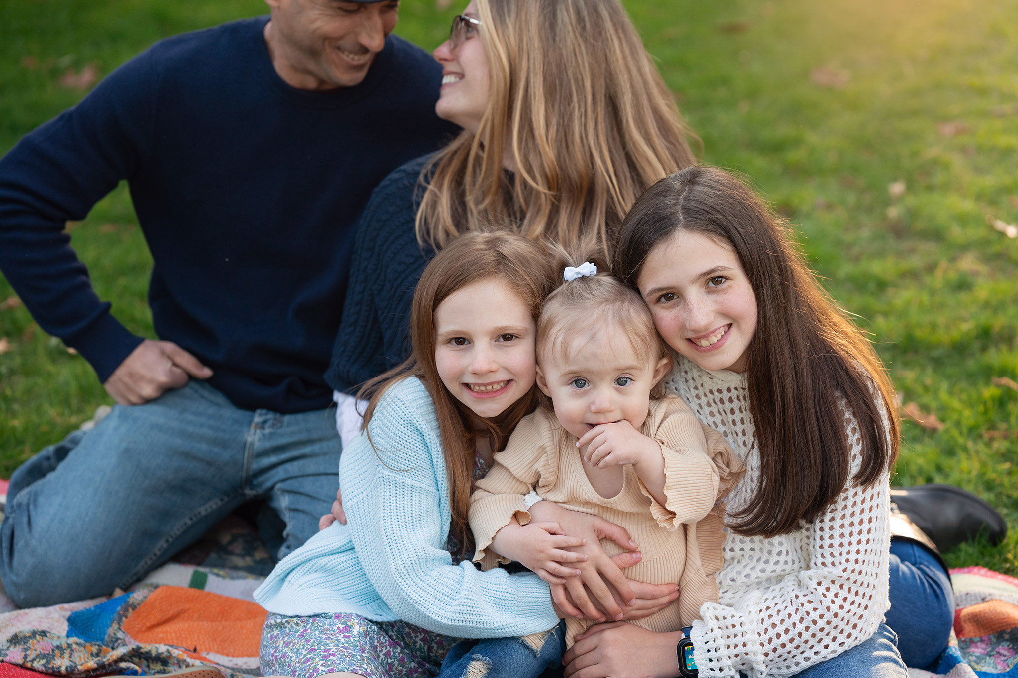 Three young sisters hug while sitting on a picnic blanket with mom and dad at sunset thanks to fertility acupuncture nj