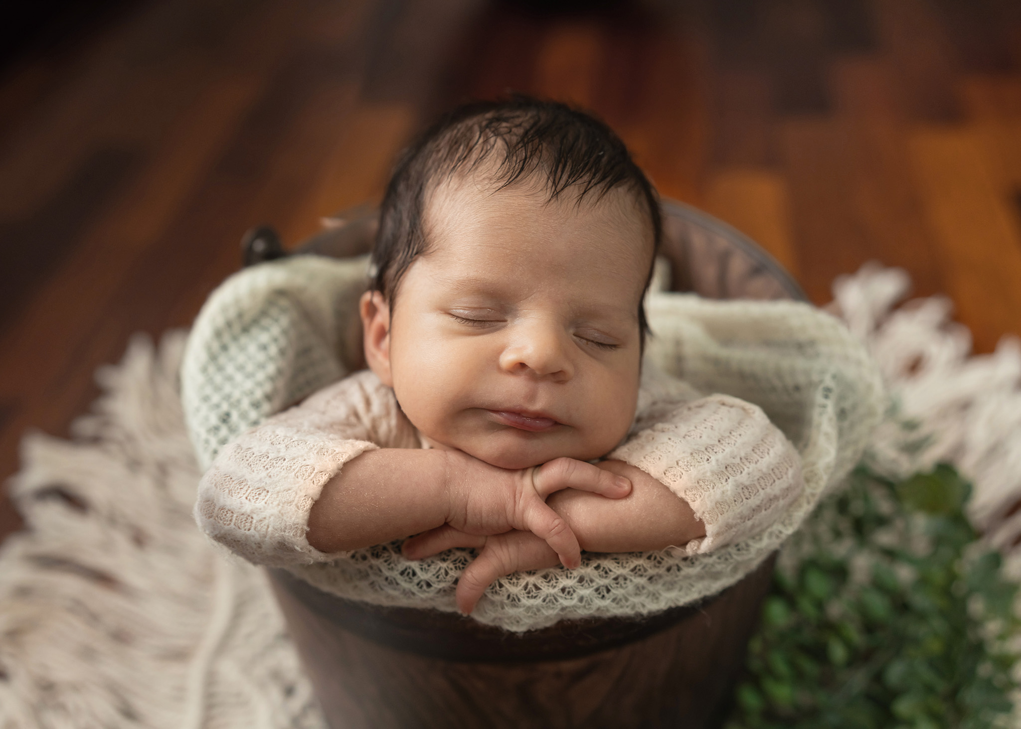 A newborn baby sleeps in a wooden bucket with head resting on hands