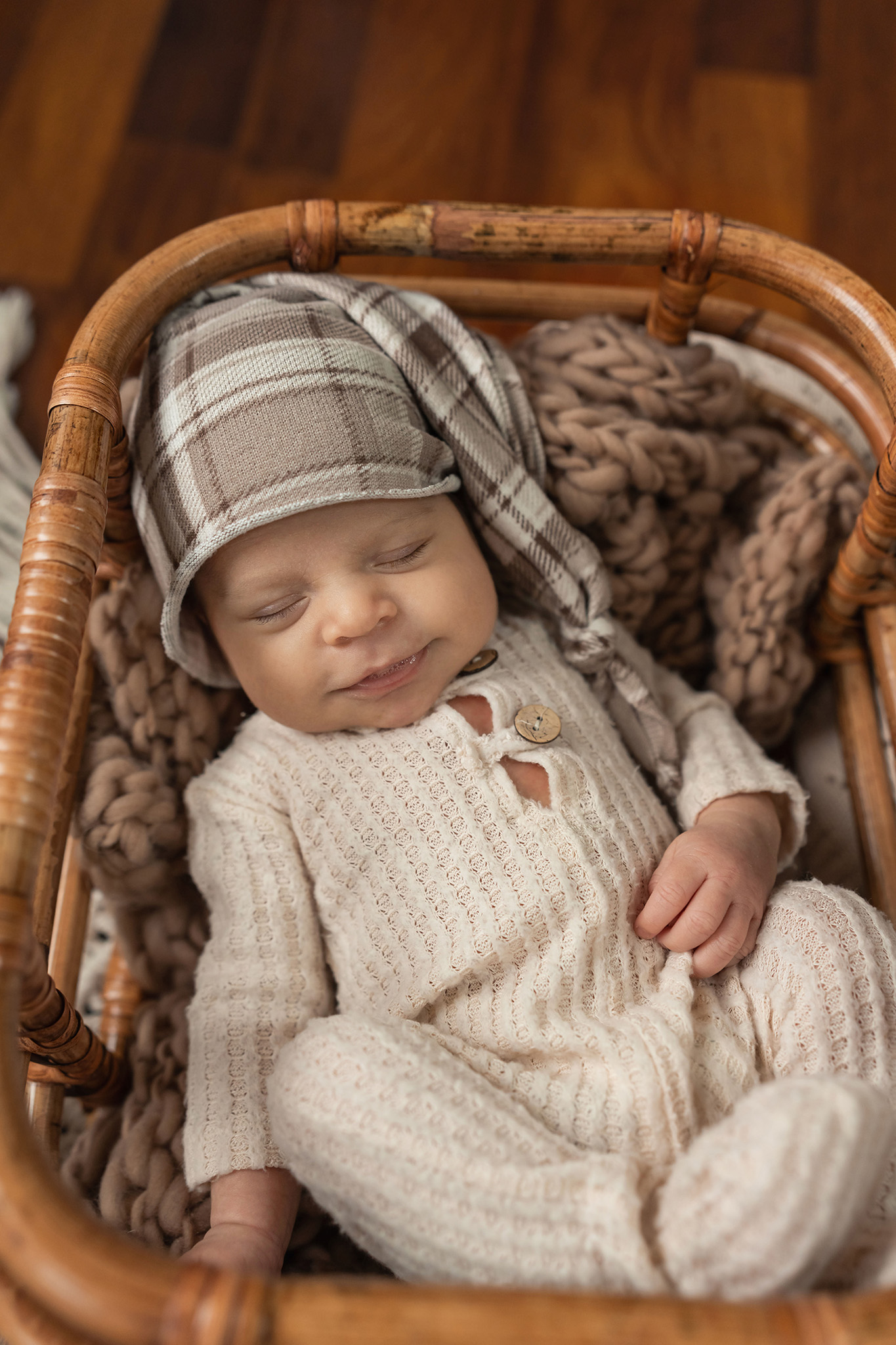 A newborn baby smiles in his sleeps while laying in a wicker basket in a plaid sleep cap