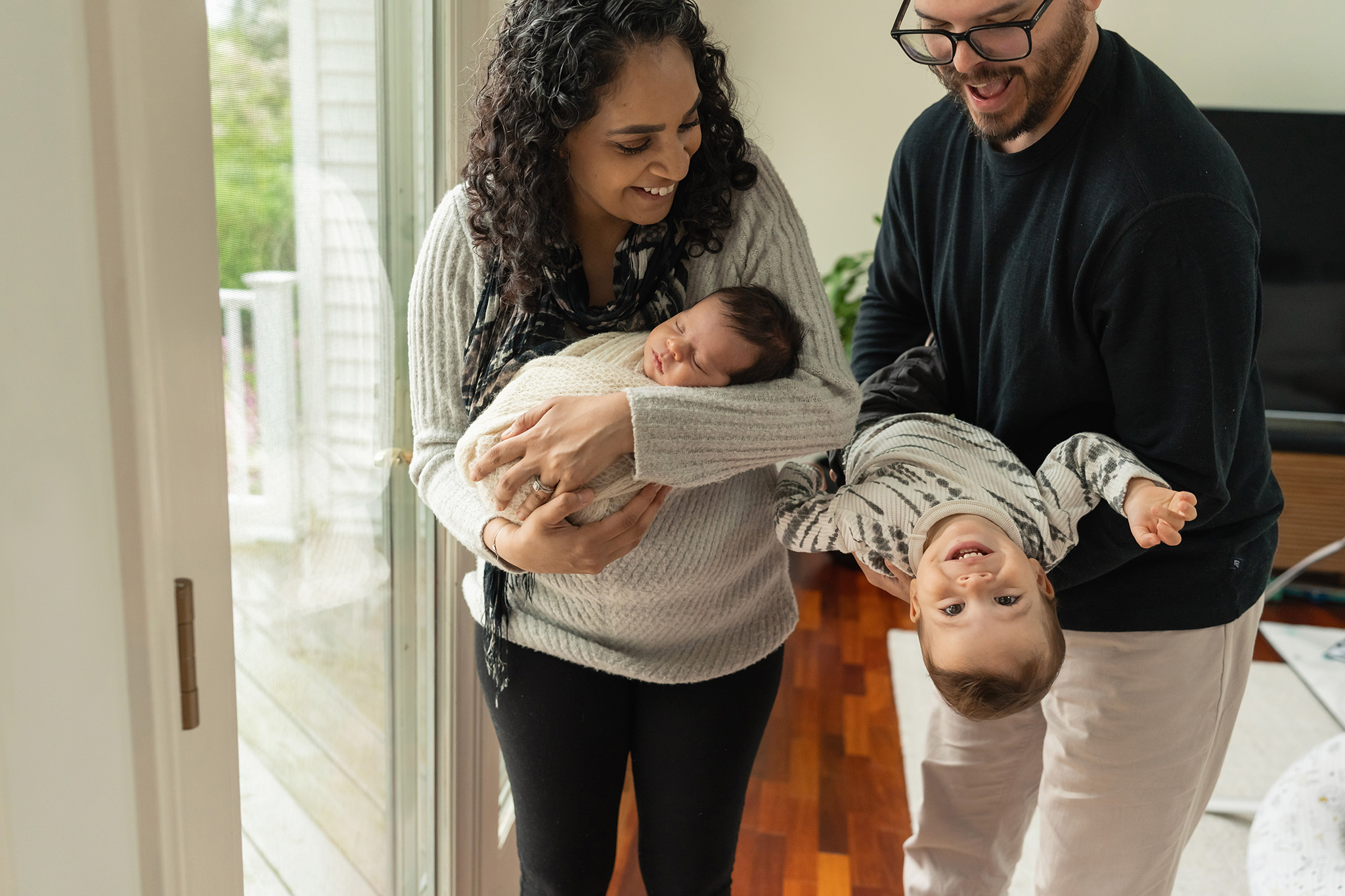 Happy parents hold and flip upside their toddler son while holding a sleeping newborn after meeting lactation consultant nj