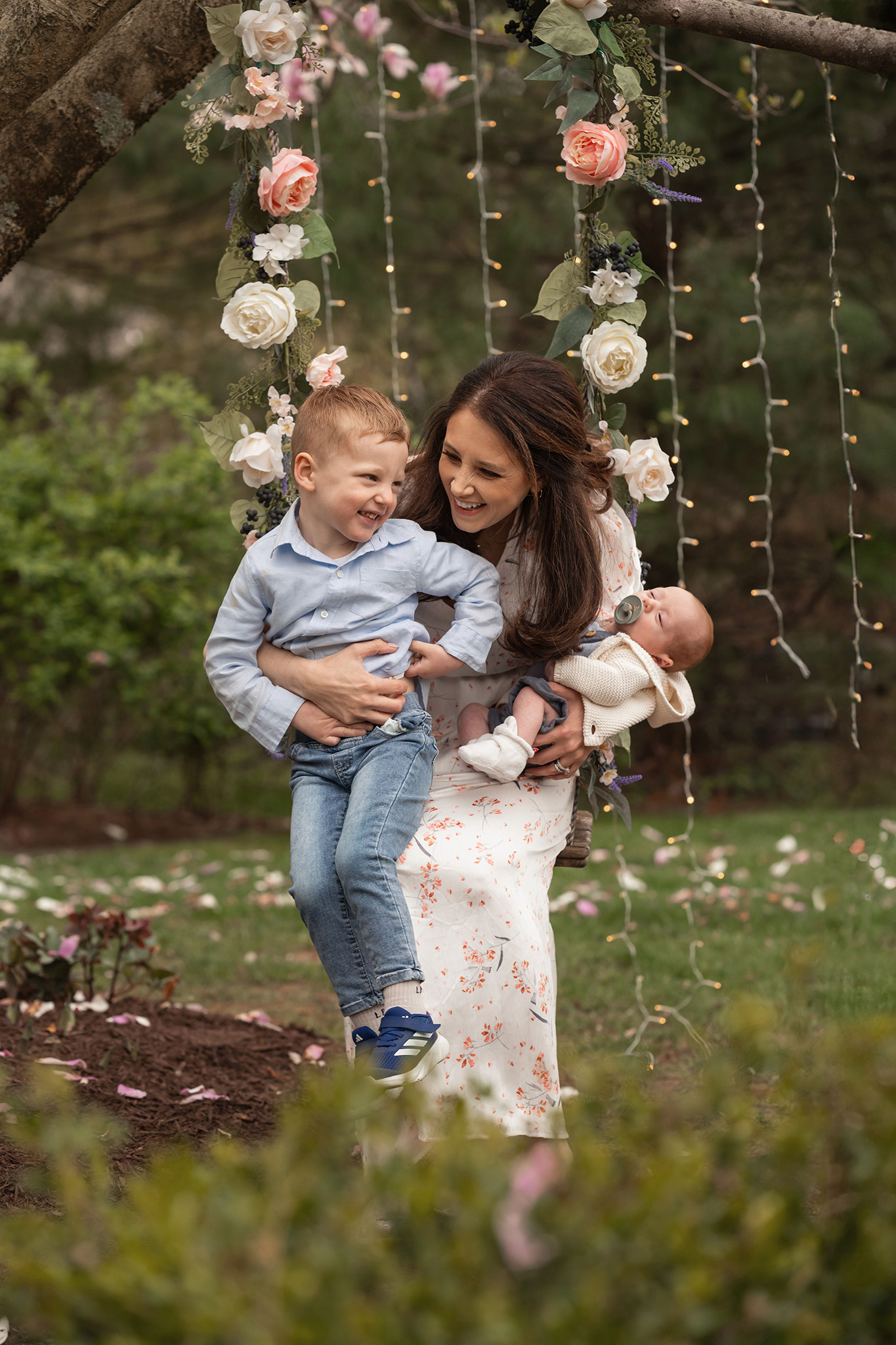 A happy mom plays with her toddler son in a garden while holding her sleeping newborn baby