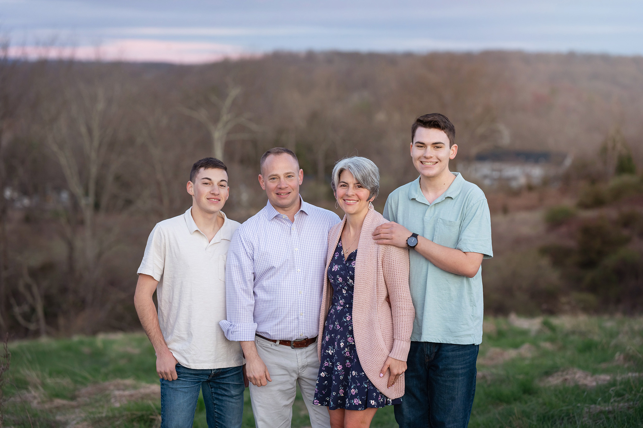 A mom and dad stand on a hill in a park with their two teenage sons at sunset