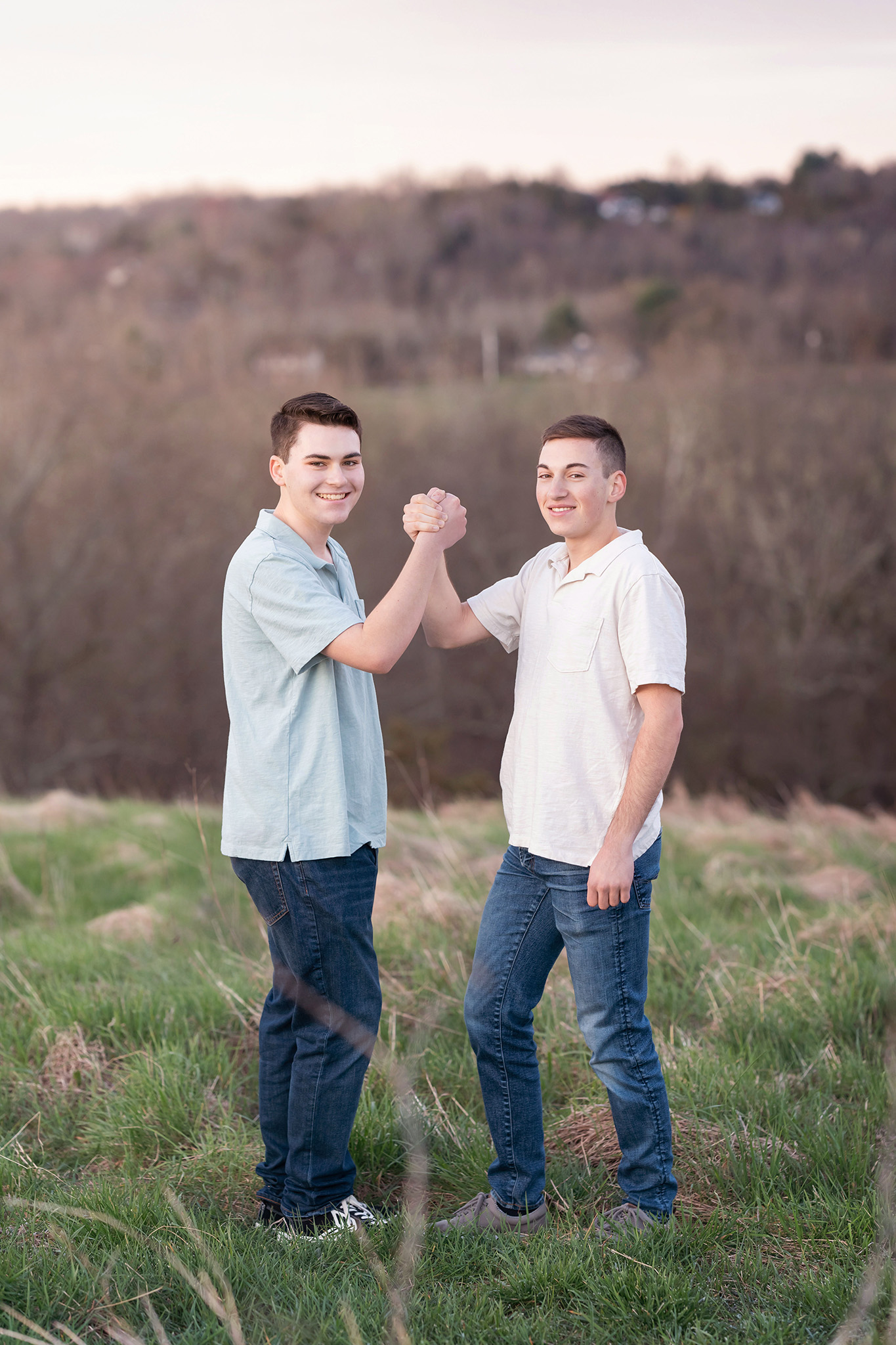 Two happy brothers lock hands while standing on a hill in grass at sunset