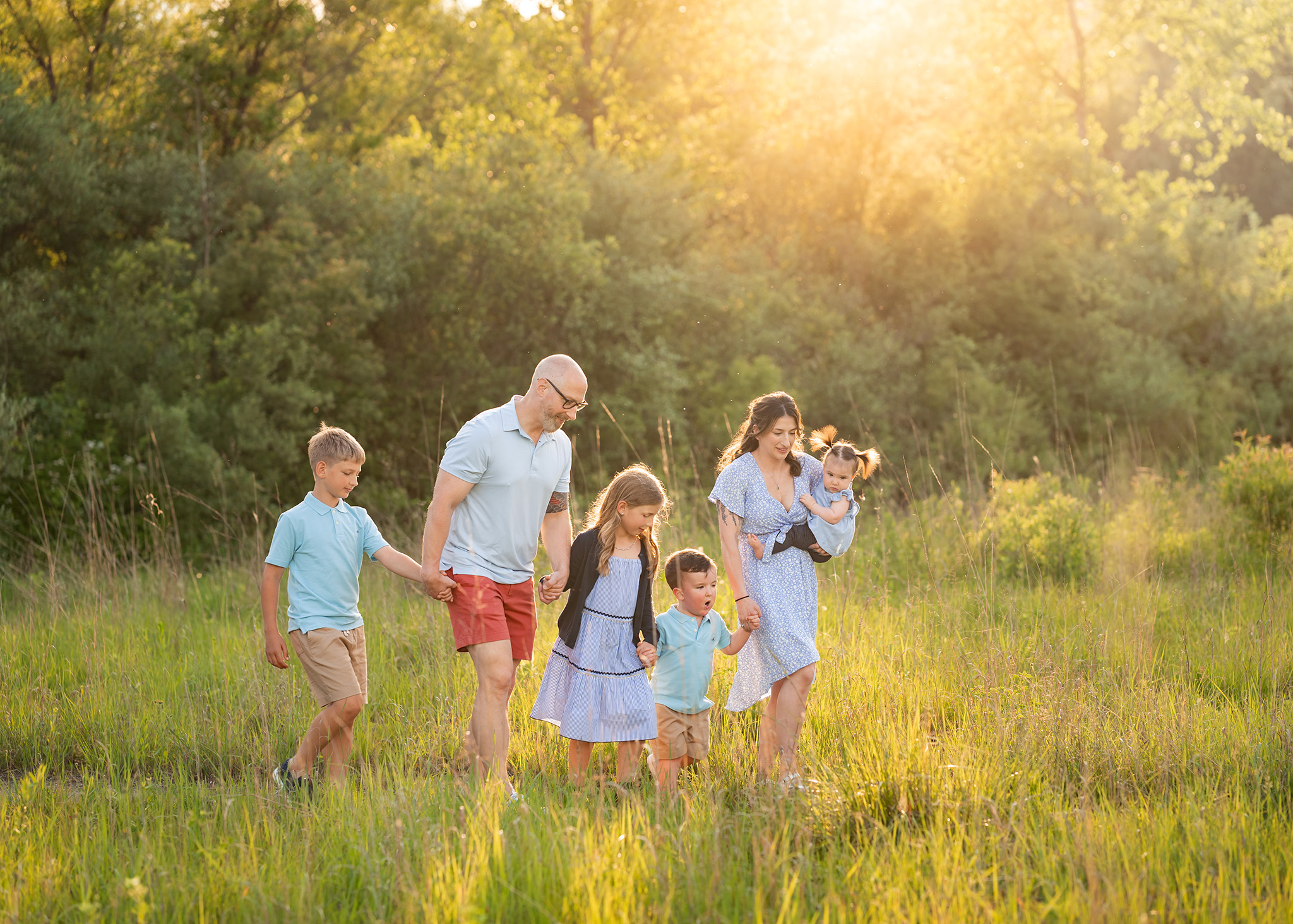 A family of six in blue shirts and dresses walk through a park trail at sunset while holding hands