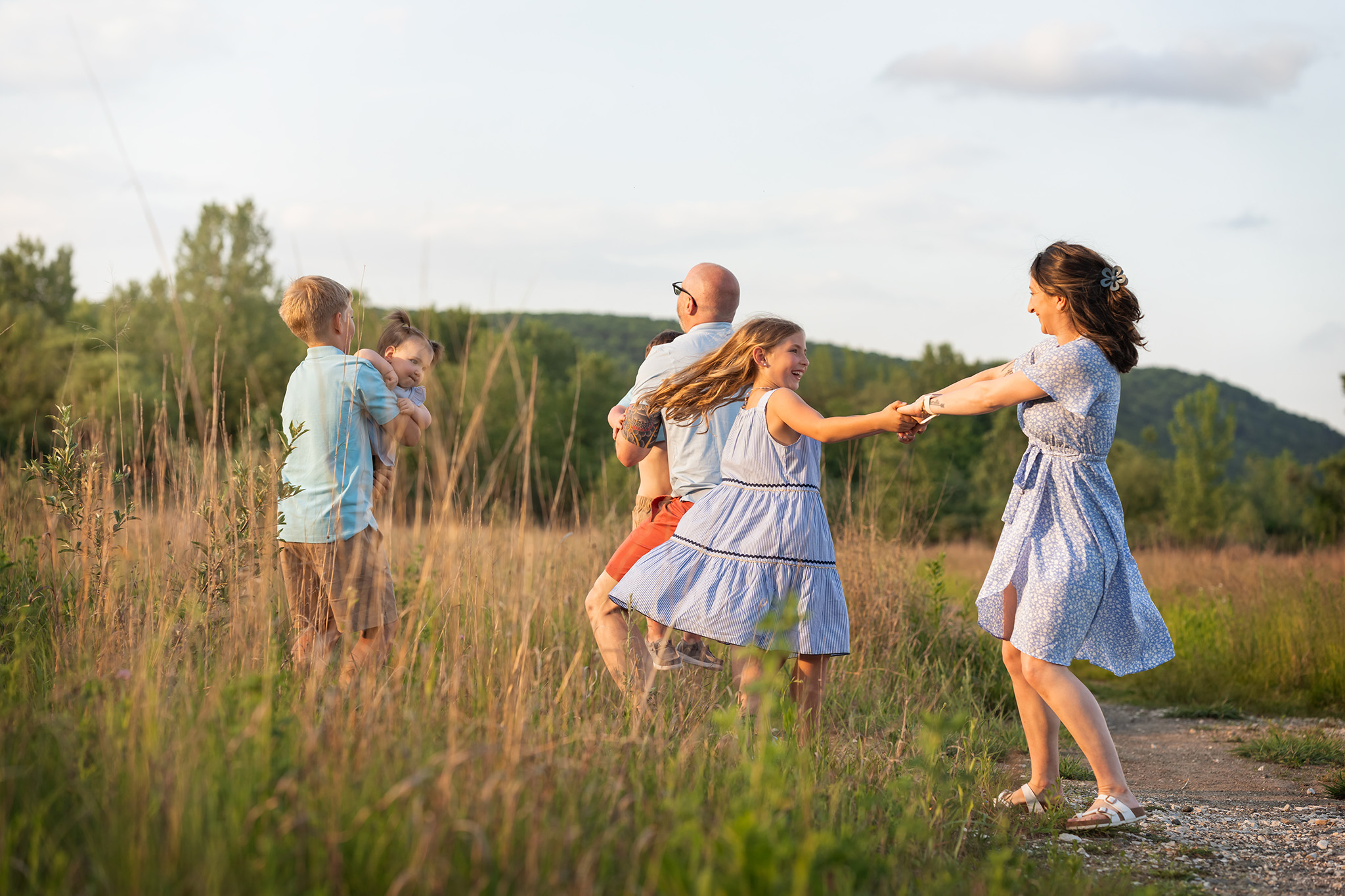 A mother and young daughter dance in a field at sunset with the rest of their family behind them