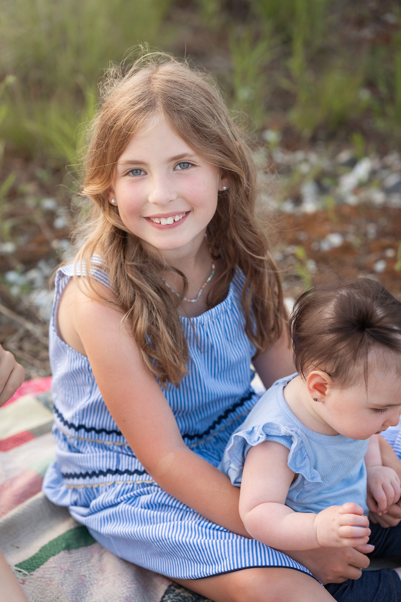 A happy toddler girl sits on a picnic blanket with her baby sister in her lap after visiting a pediatric dentist northern nj