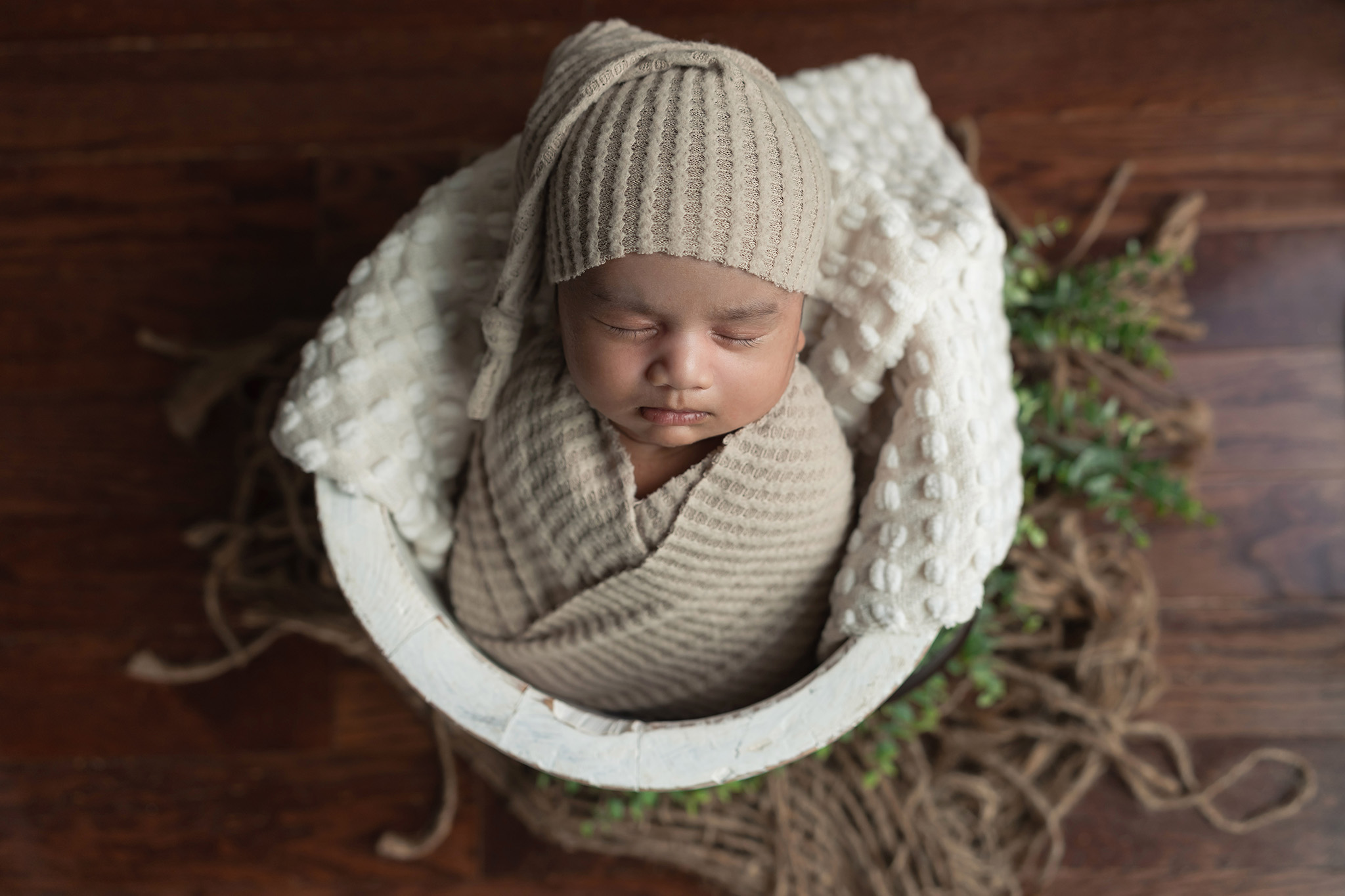 A newborn baby sleeps in a tan sleep cap and matching swaddle in a wooden bucket