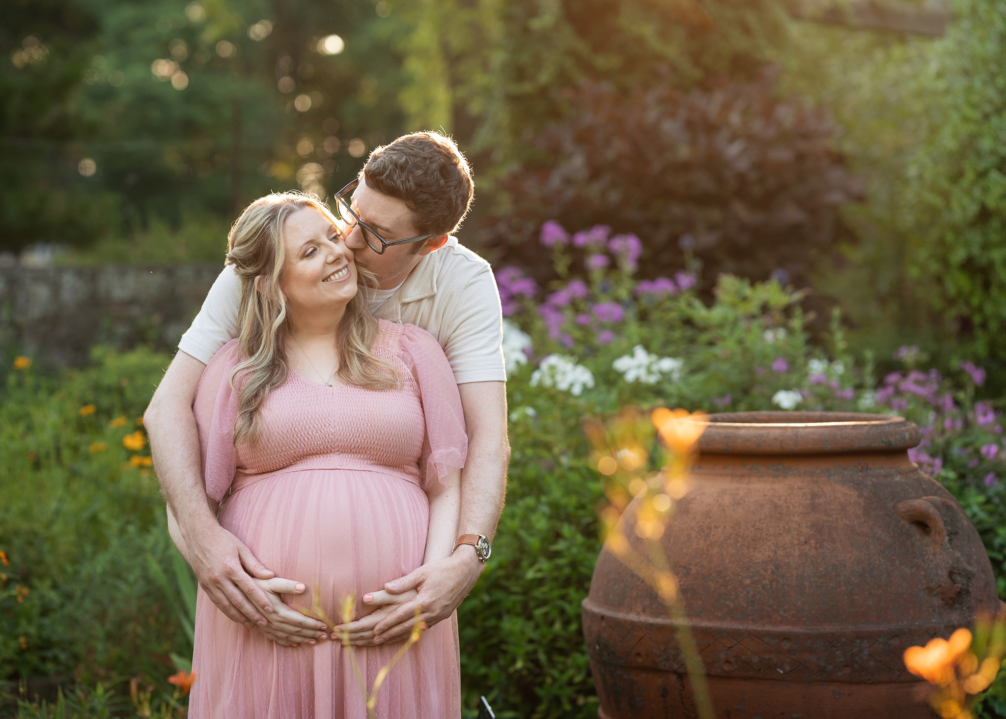 A mom to be in a pink maternity gown smiles big while being kissed on the cheek in a garden at sunset before finding placenta encapsulation nj