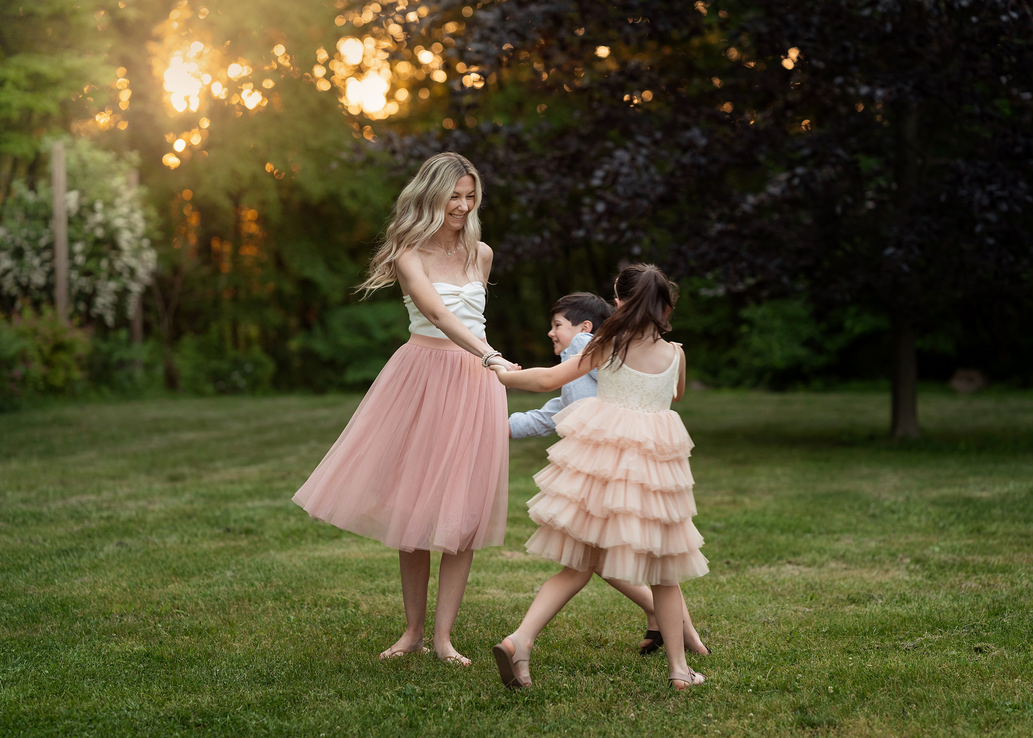 A happy mom in a pink dress dances with her young son and daughter in a park at sunset thanks to help from prenatal yoga morristown nj