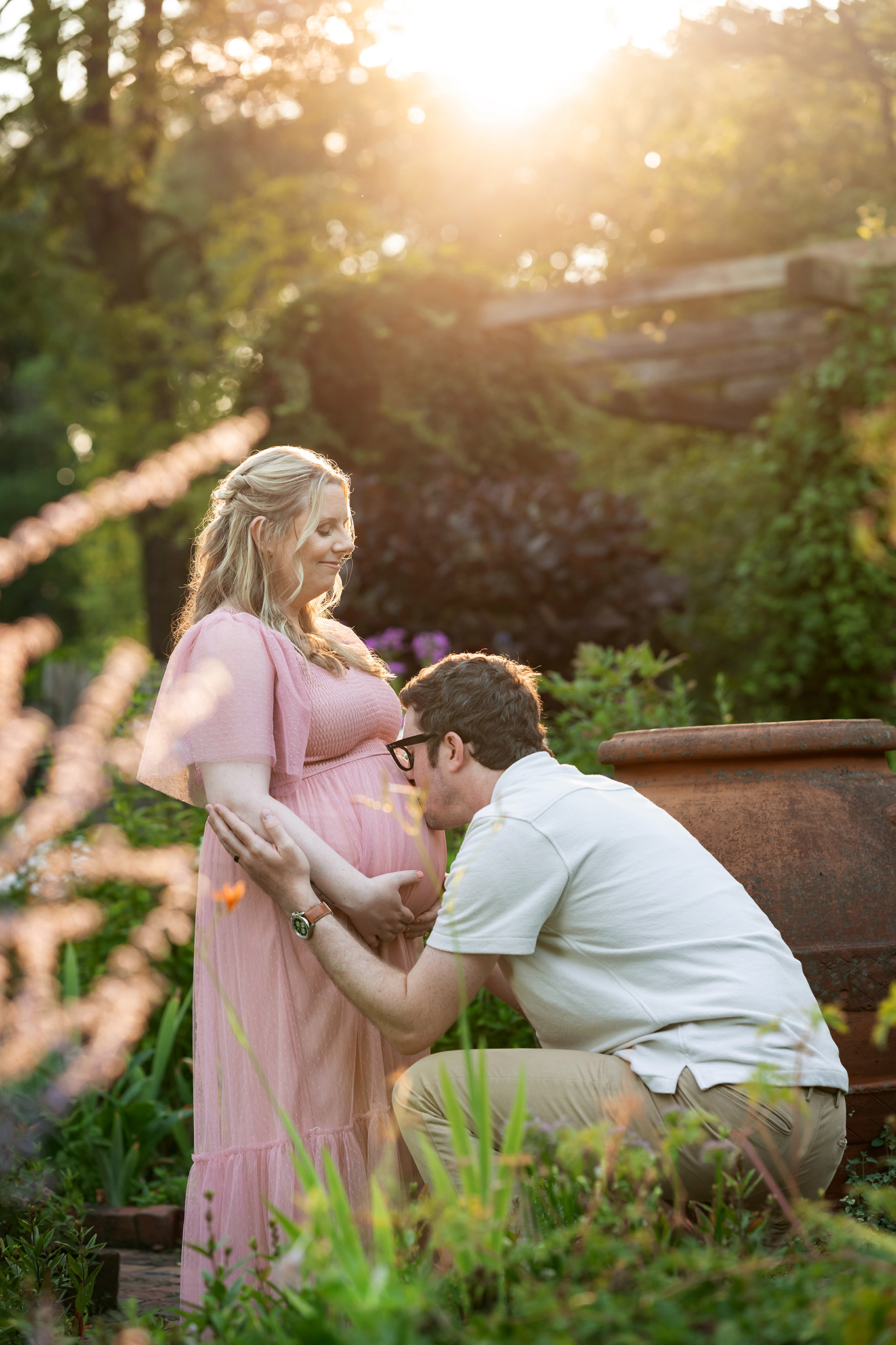 An expecting father kisses the bump of his pregnant wife in a pink maternity gown in a garden at sunset