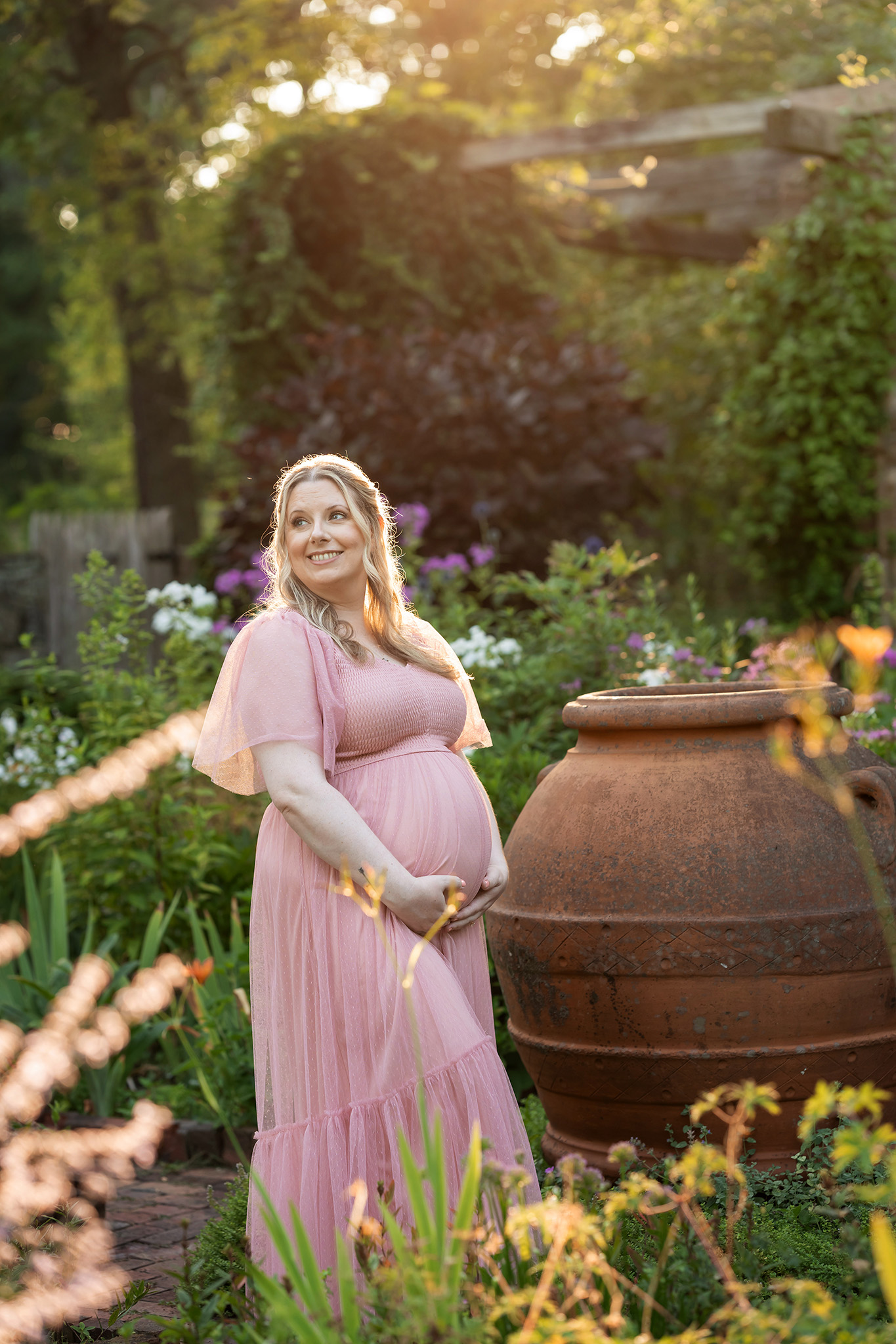 A happy mother to be in a pink maternity gown explores a garden at sunset while holging her bump after some prenatal yoga northern nj