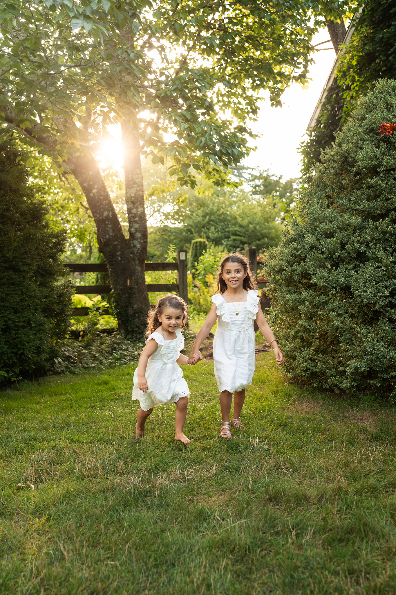 Toddler sisters in white dresses giggle while exploring a lawn at sunset and holding hands