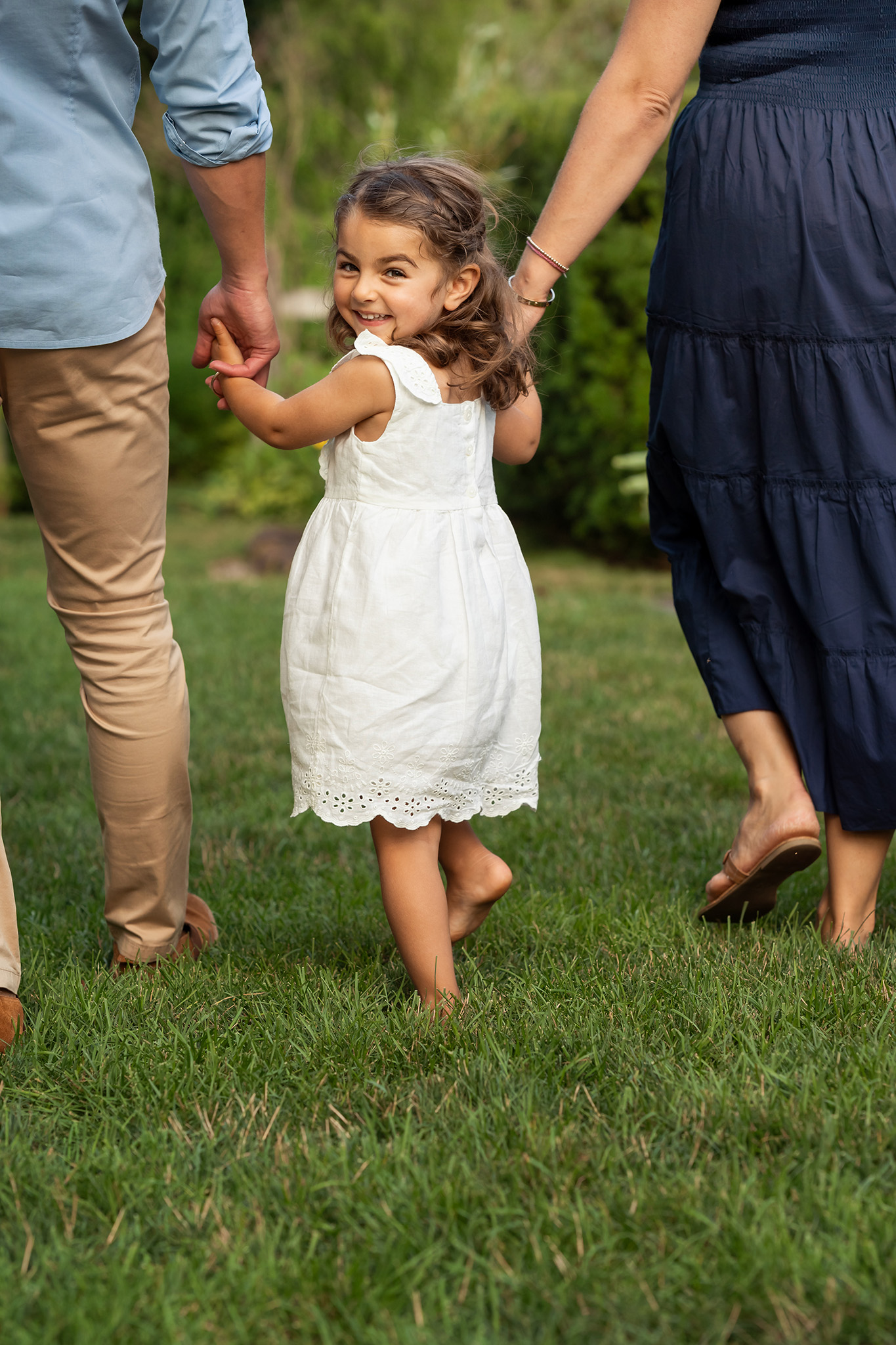 A happy toddler girl in a white dress walks in a lawn while holding hands with mom and dad before some swimming lessons northern nj