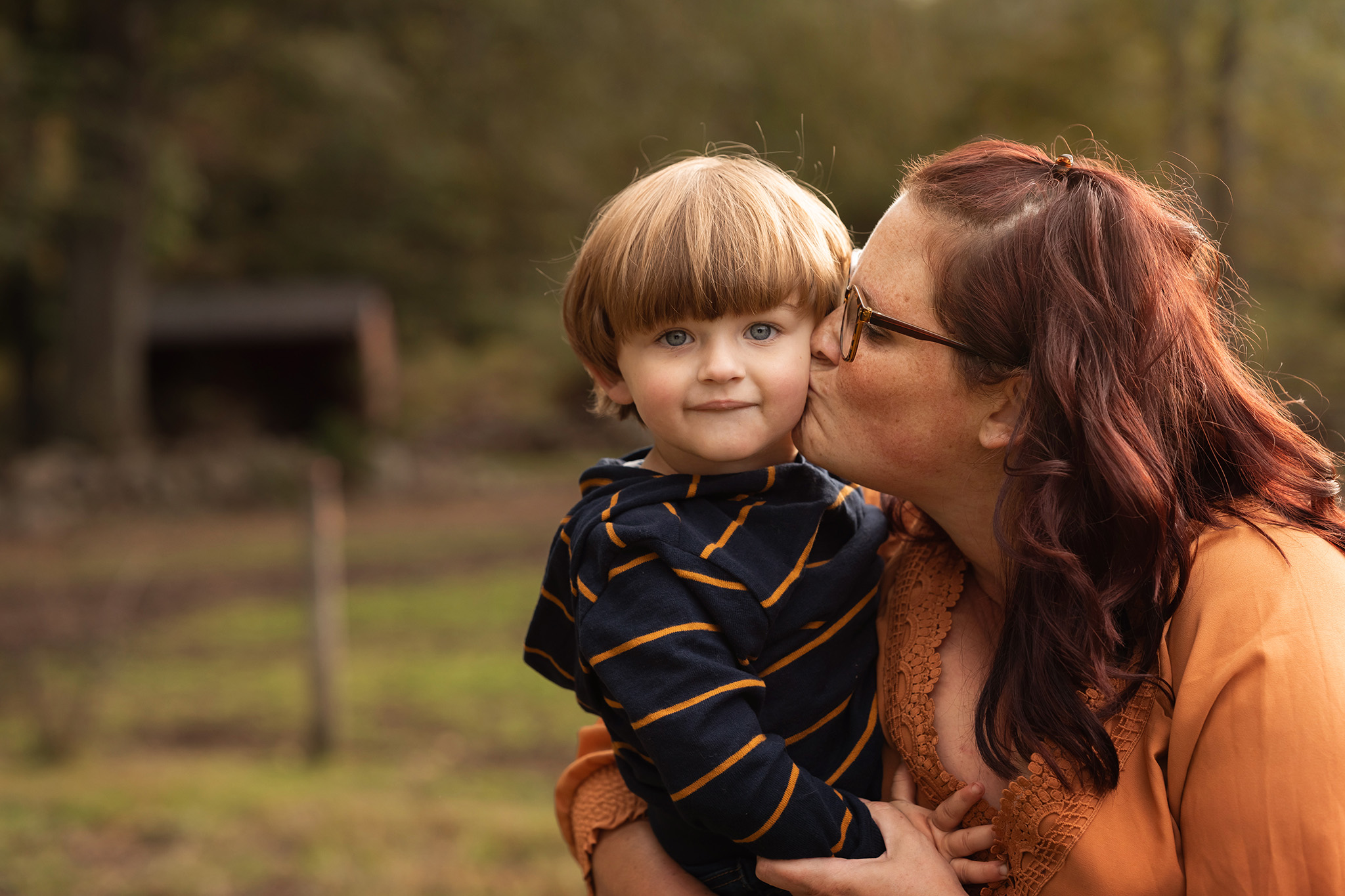 A toddler boy in a stripe shirt is kissed on the cheek by mom while sitting in her arms