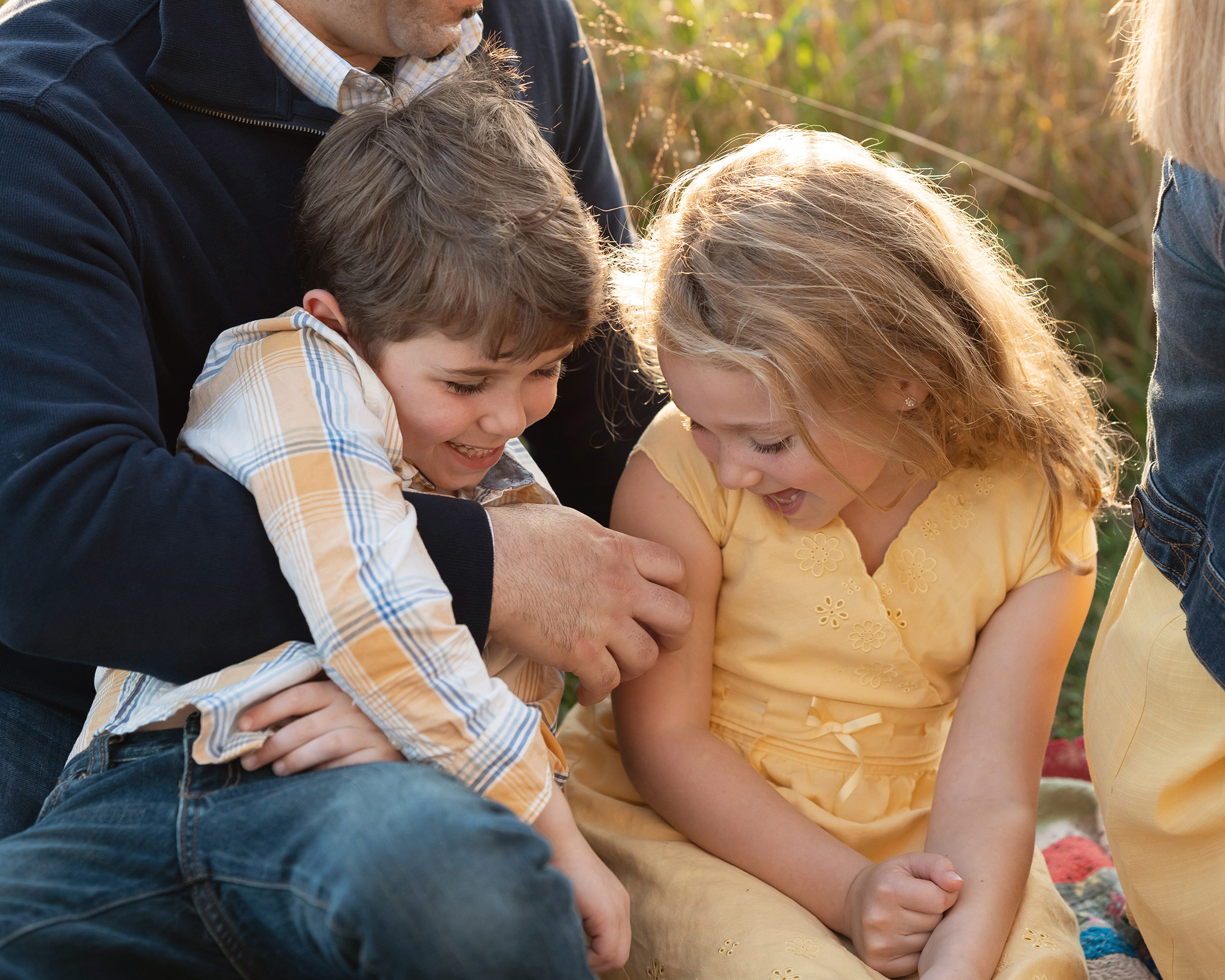 Toddler brother and sister giggle while being tickled in dad's lap in a park field before visiting toy stores nj