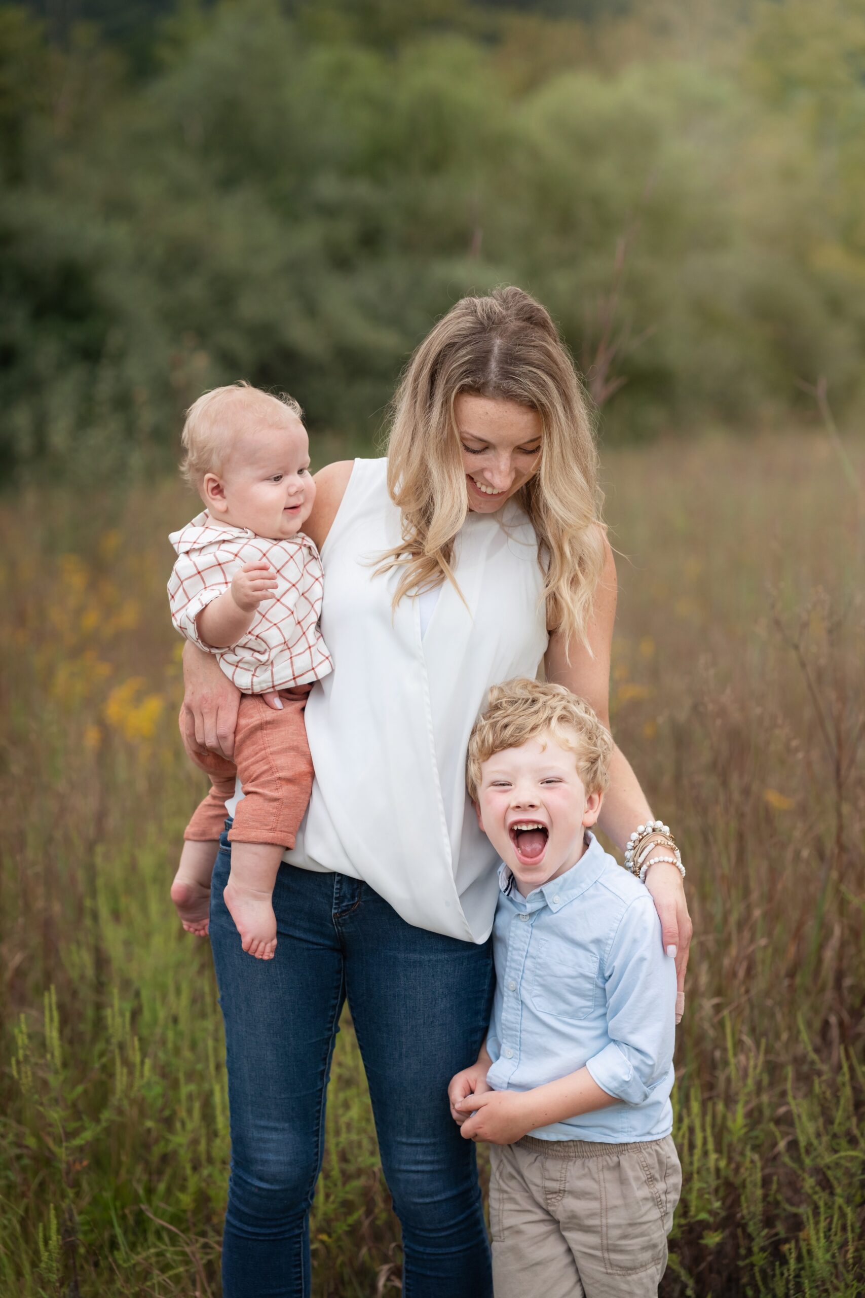 A mom walks in tall grass with a baby on her hip and toddler laughing at her side in jeans and white top