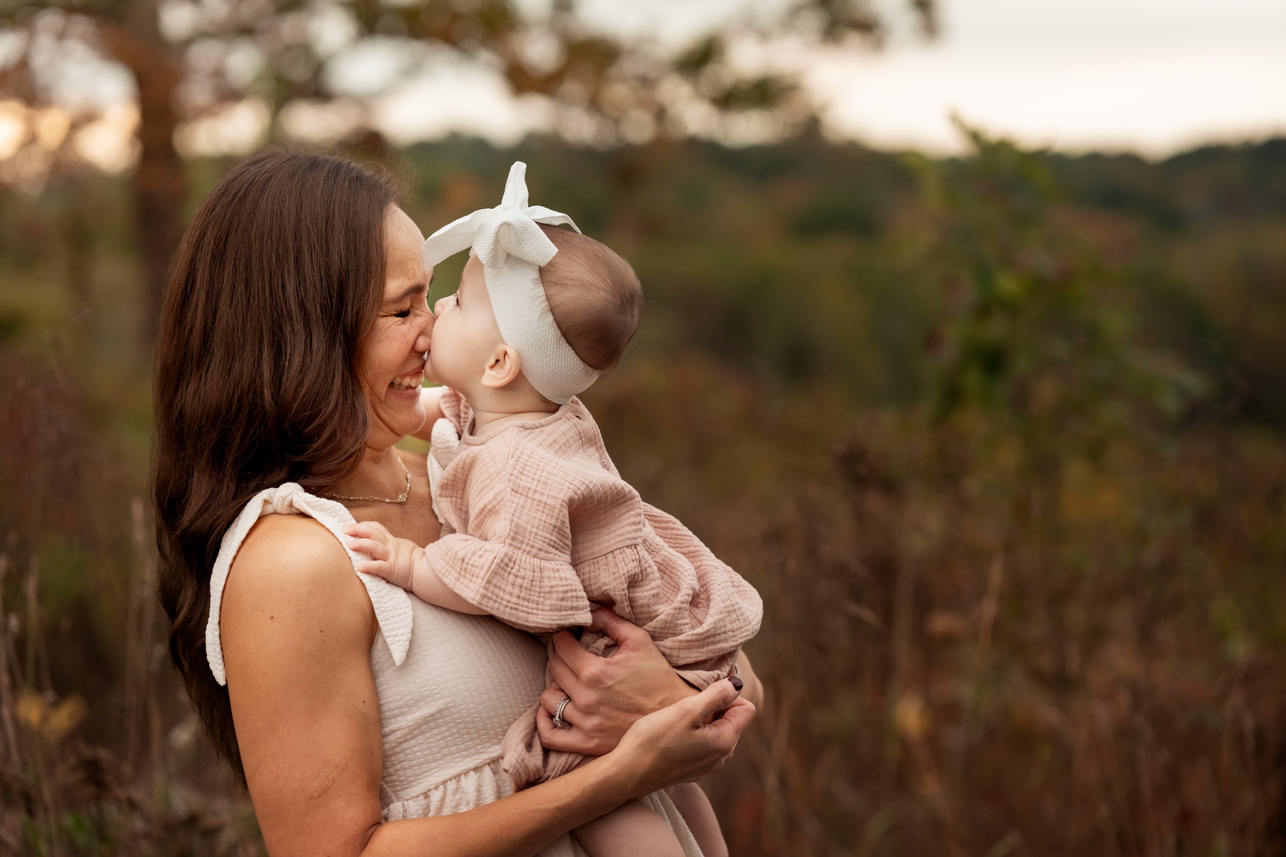 A laughing mom has her nose smushed by her baby daughter in her arms in a pink dress