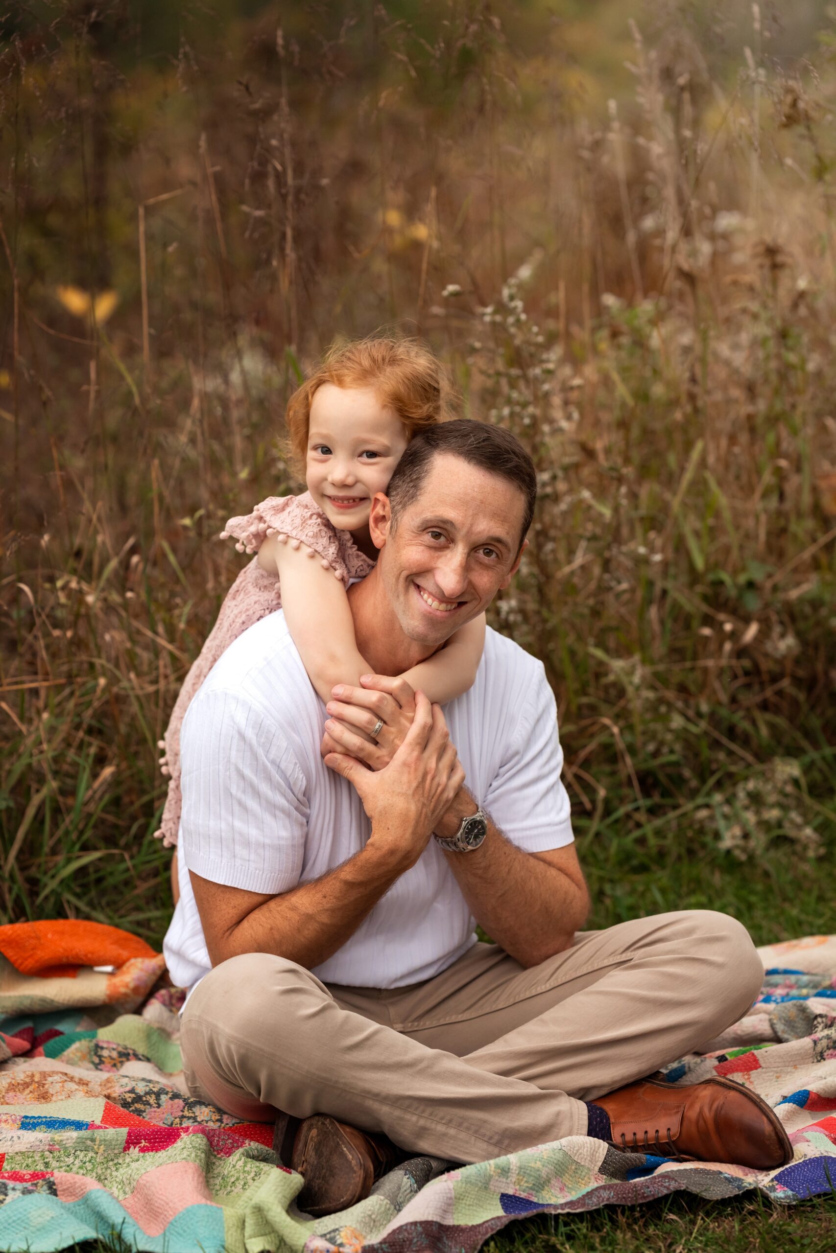 A toddler girl in a pink dress hugs dad's neck from behind while on a picnic blanket at sunset after visiting pediatricians in cranford nj