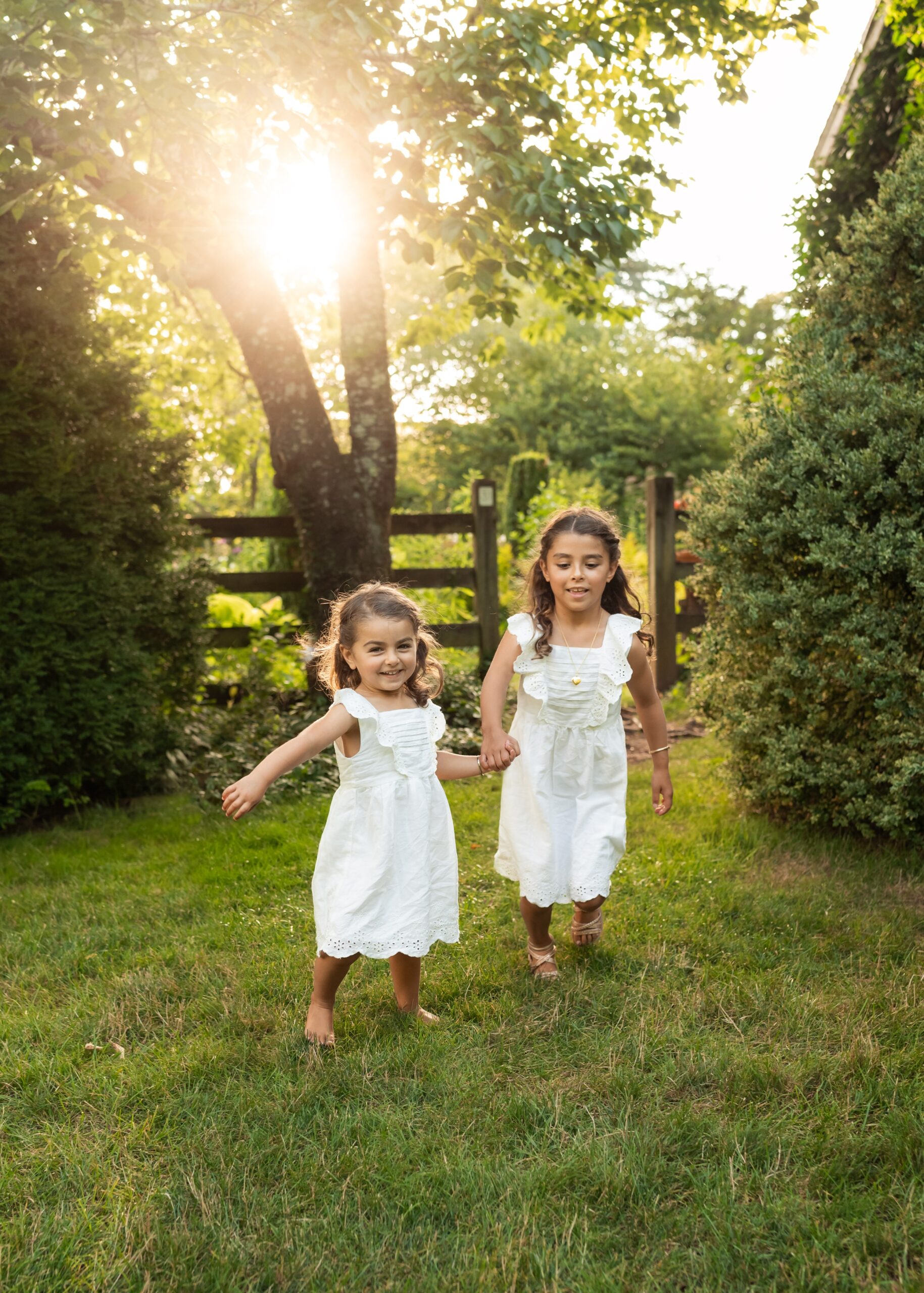 Two toddler sisters in white dresses hold hadns and explore a garden at sunset