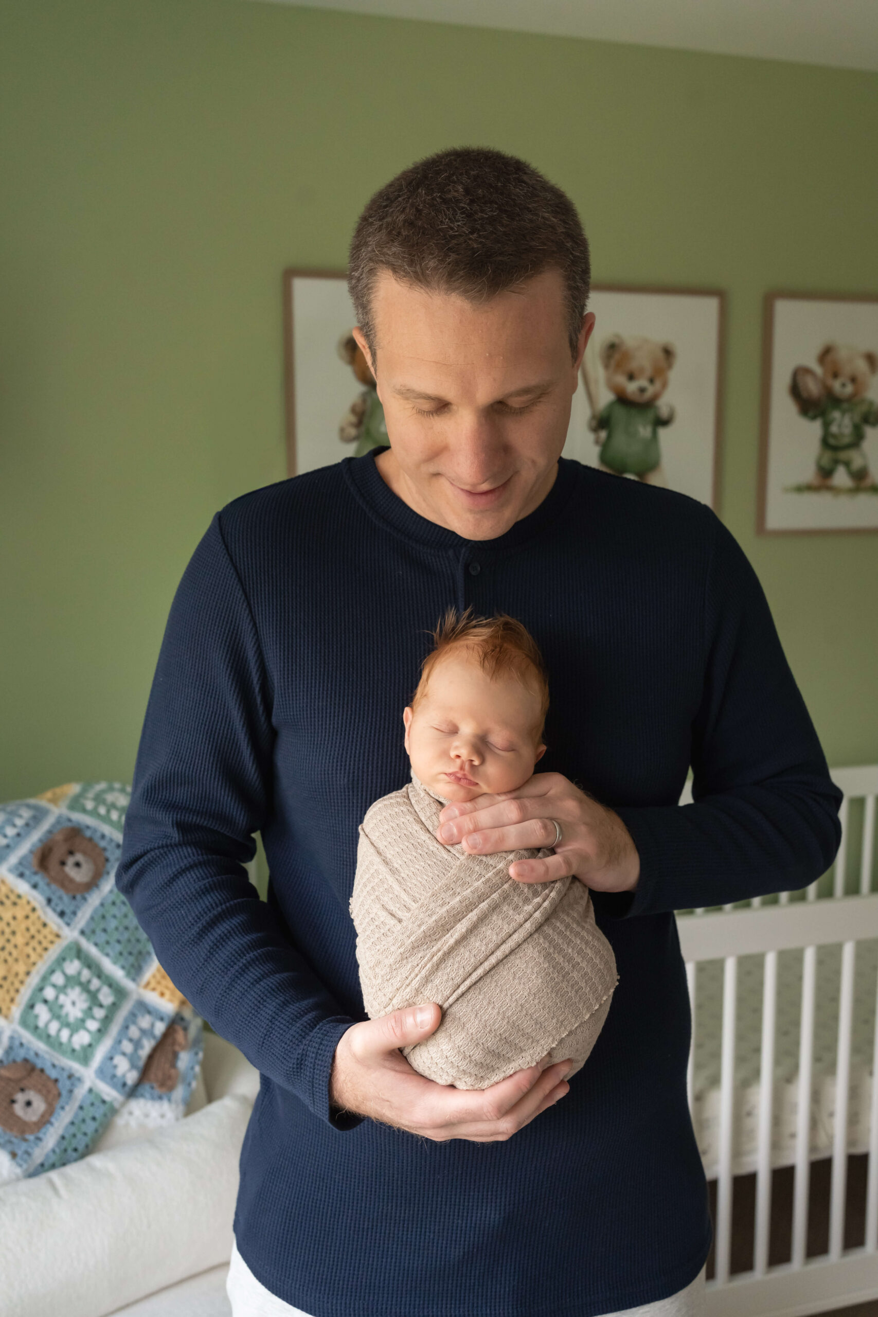 A newborn baby in a tan swaddle sleeps while leaning against dad's chest in a nursery