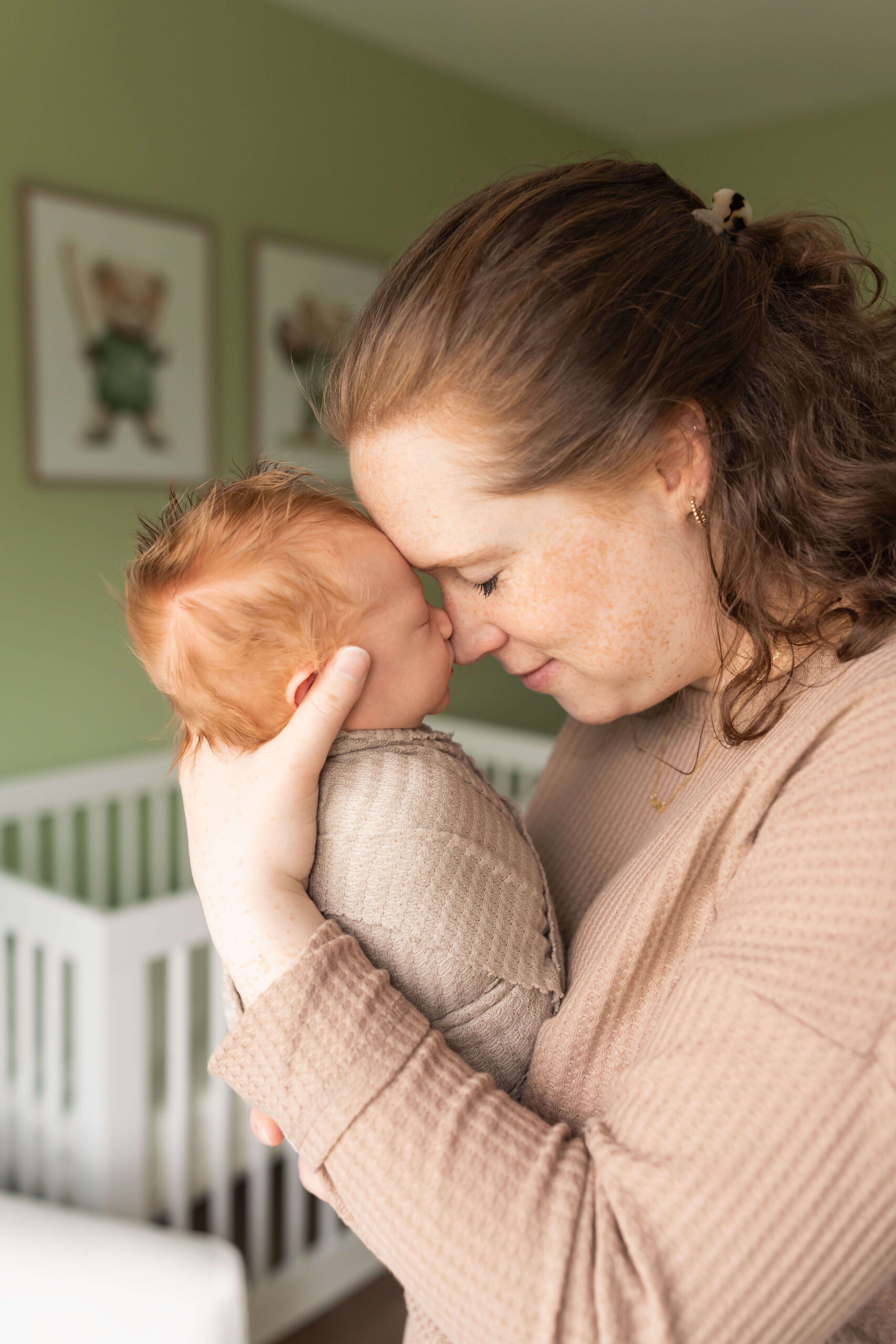 A new mom touches foreheads and noses with her sleeping newborn while standing in a nursery window in a tan sweater after meeting with beyond birth