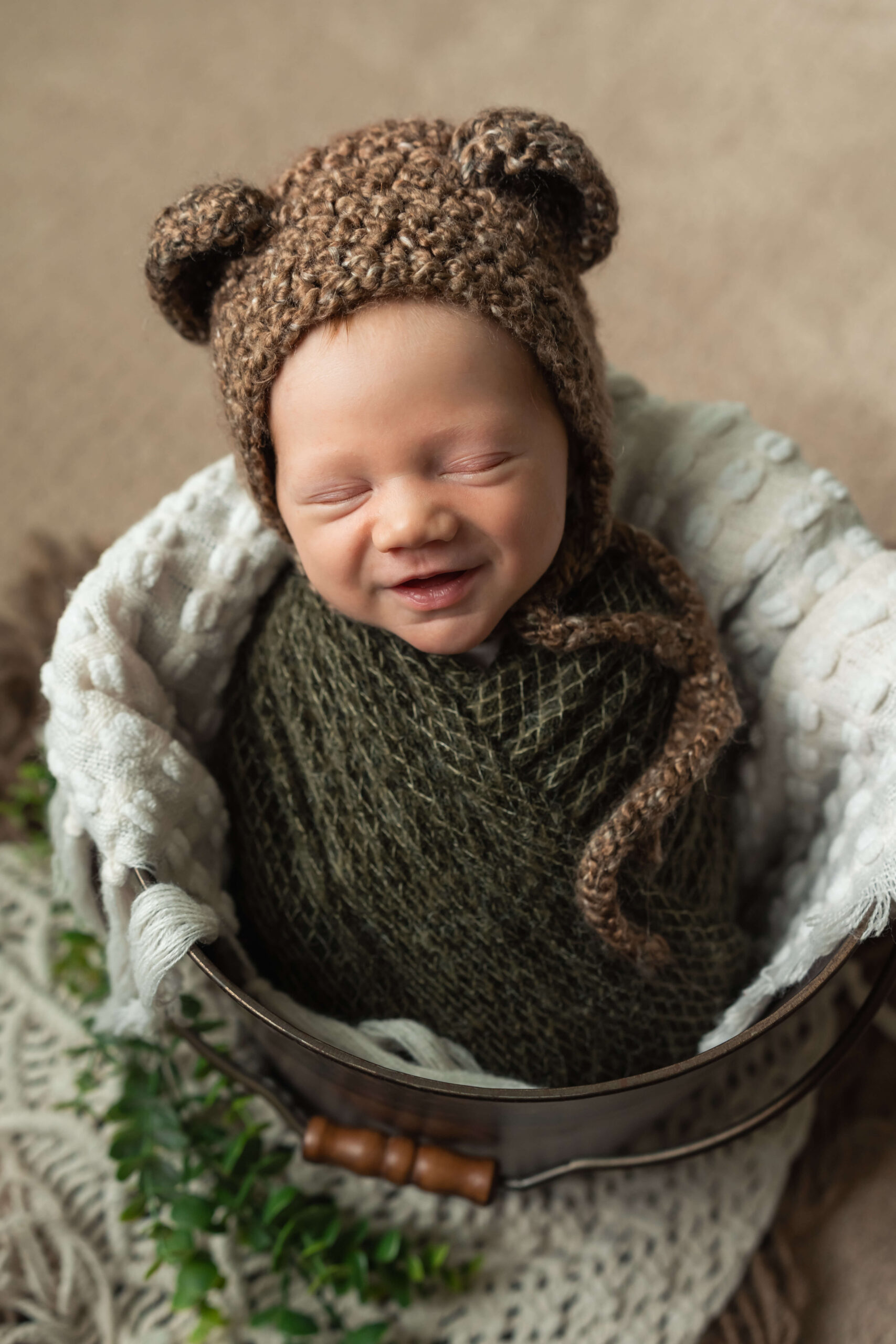 A smiling newborn sleeps in a knit bear ear bonnet in a metal bucket after meeting beyond birth