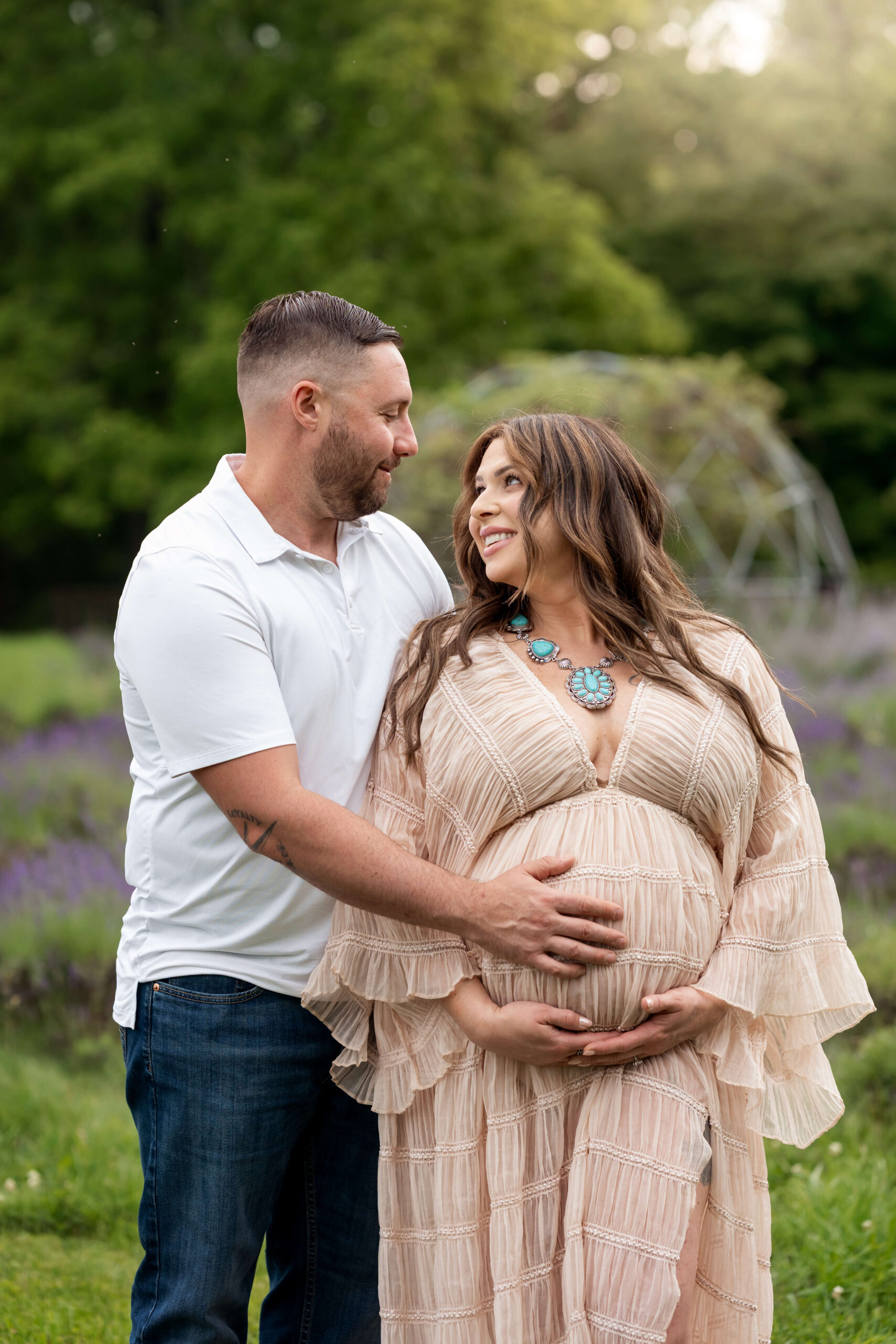 A happy expectant couple stand holding the bump and smiling at each other in a lavender garden at sunset
