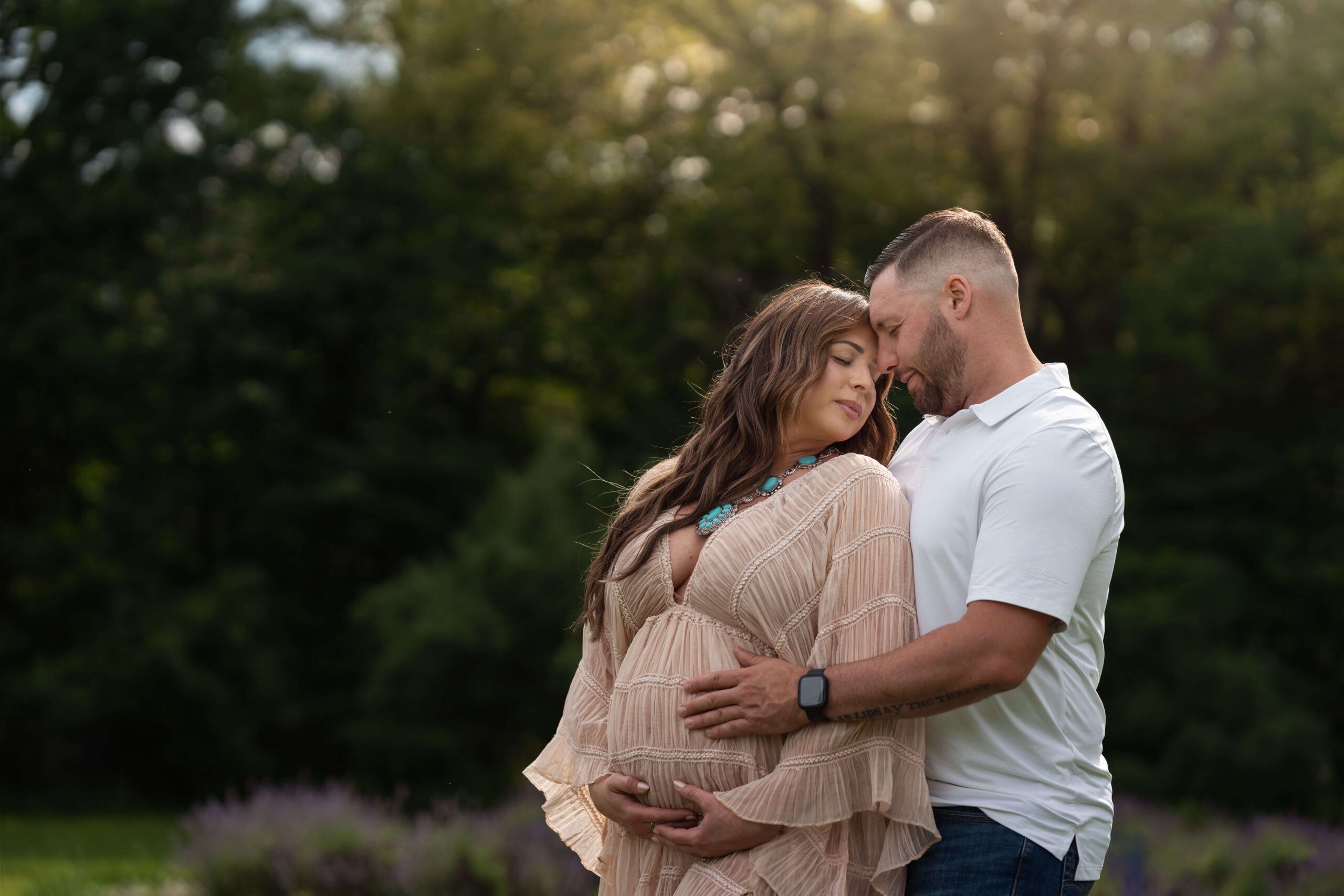 A pregnant woman leans into the chest and snuggles of her husband while wearing a tan maternity gown in a park at sunset